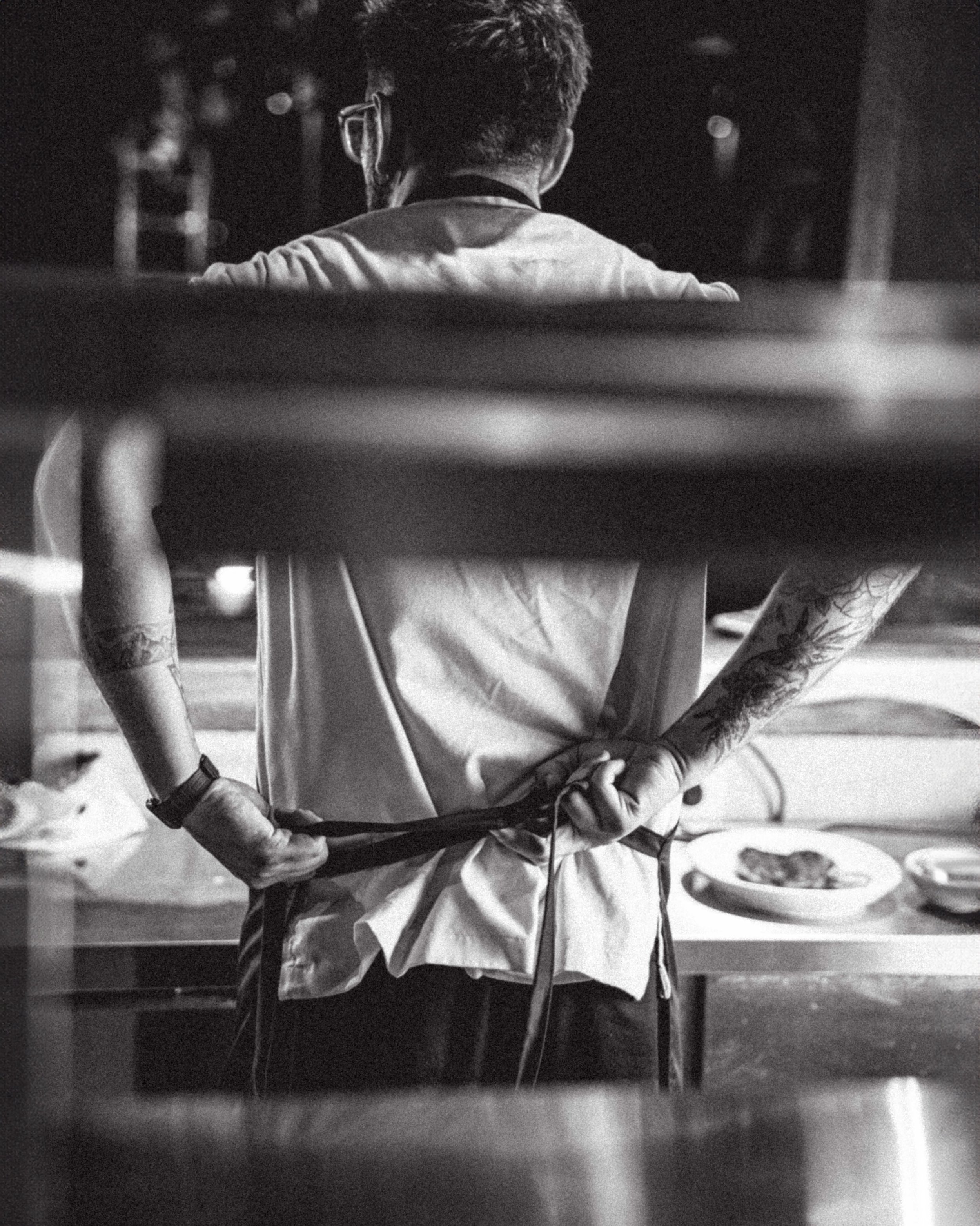 A black-and-white photo of a person with short hair, wearing glasses, a light-colored shirt, and tattoos on their arms, sitting with hands clasped while holding a smartphone, in a setting that appears to be indoors, possibly a restaurant or cafe.
