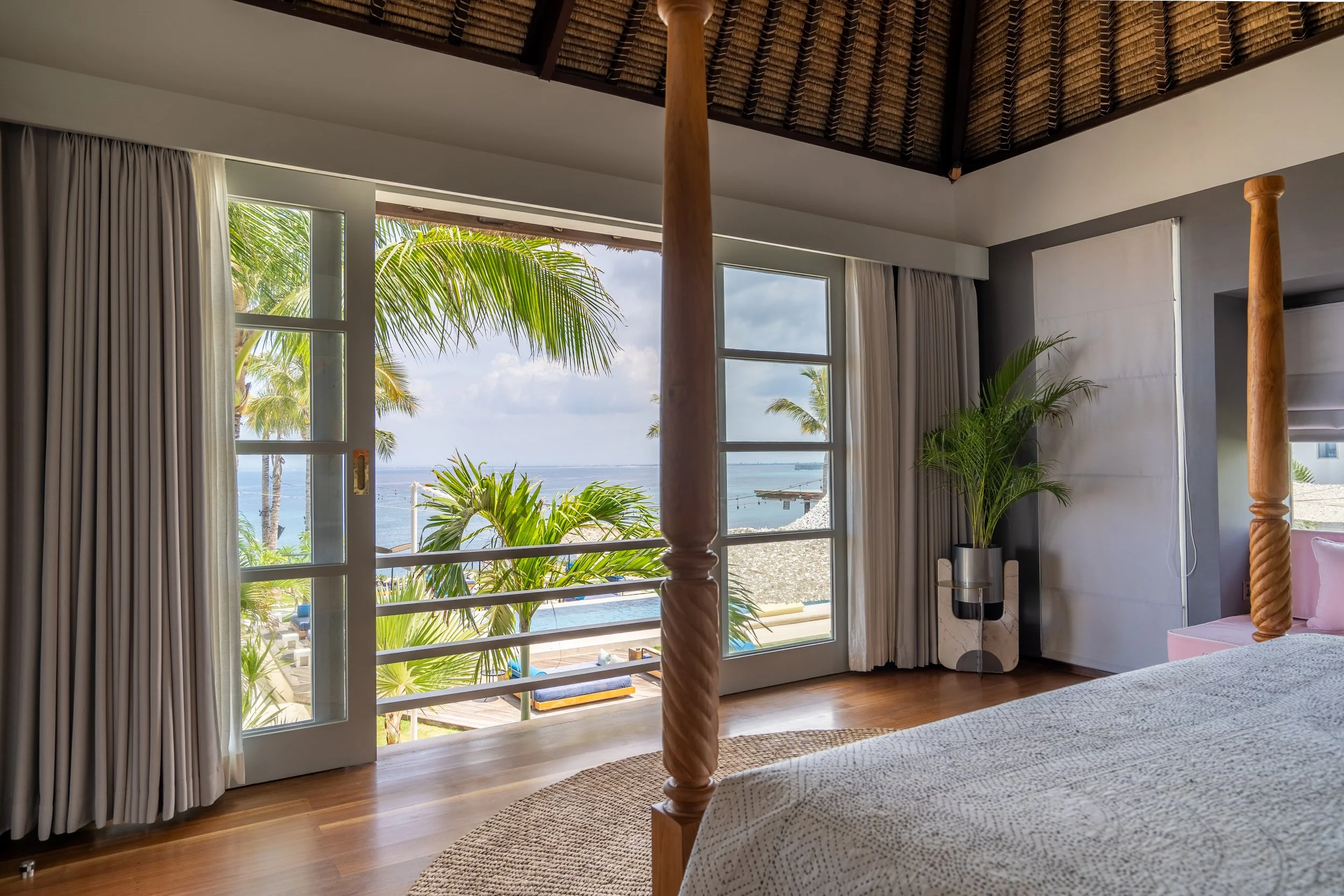 A bedroom view showing large windows with white curtains, overlooking a tropical landscape with palm trees and water. The room has wooden flooring, a canopy bed with braided posts, and a potted plant.