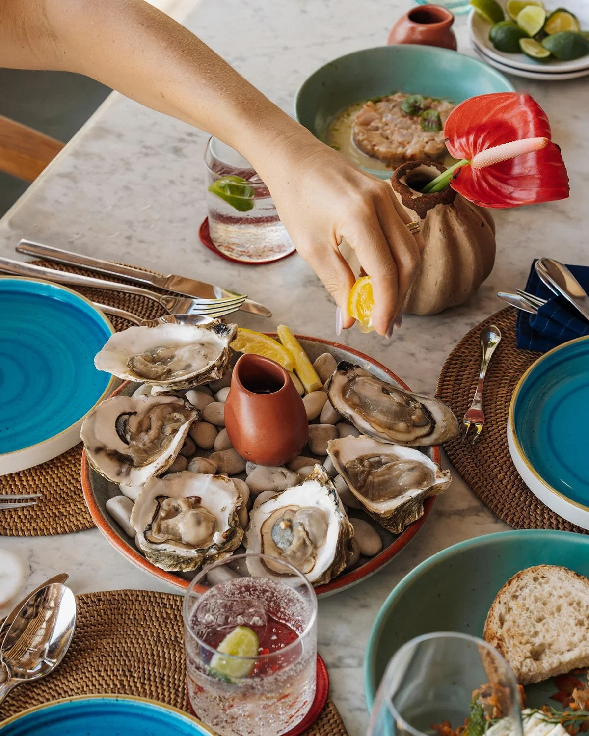 A person squeezing lemon over fresh oysters on a bed of rocks, with a variety of colorful dishes and drinks on a marble table.