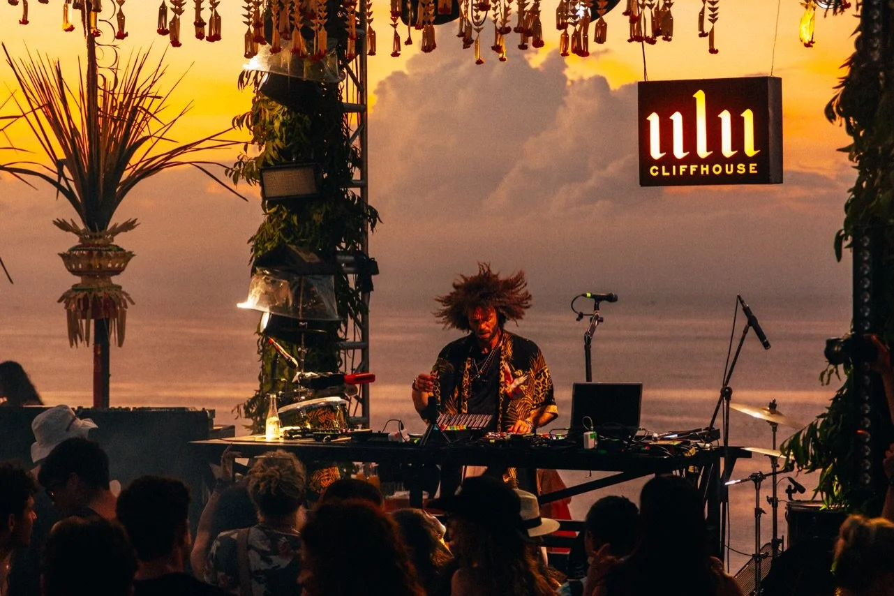 A DJ performs on stage at a seaside venue during sunset, with an audience watching. The stage is decorated with tropical plants and hanging ornaments, and there is a sign reading 'ullu CLIFFHOUSE'.