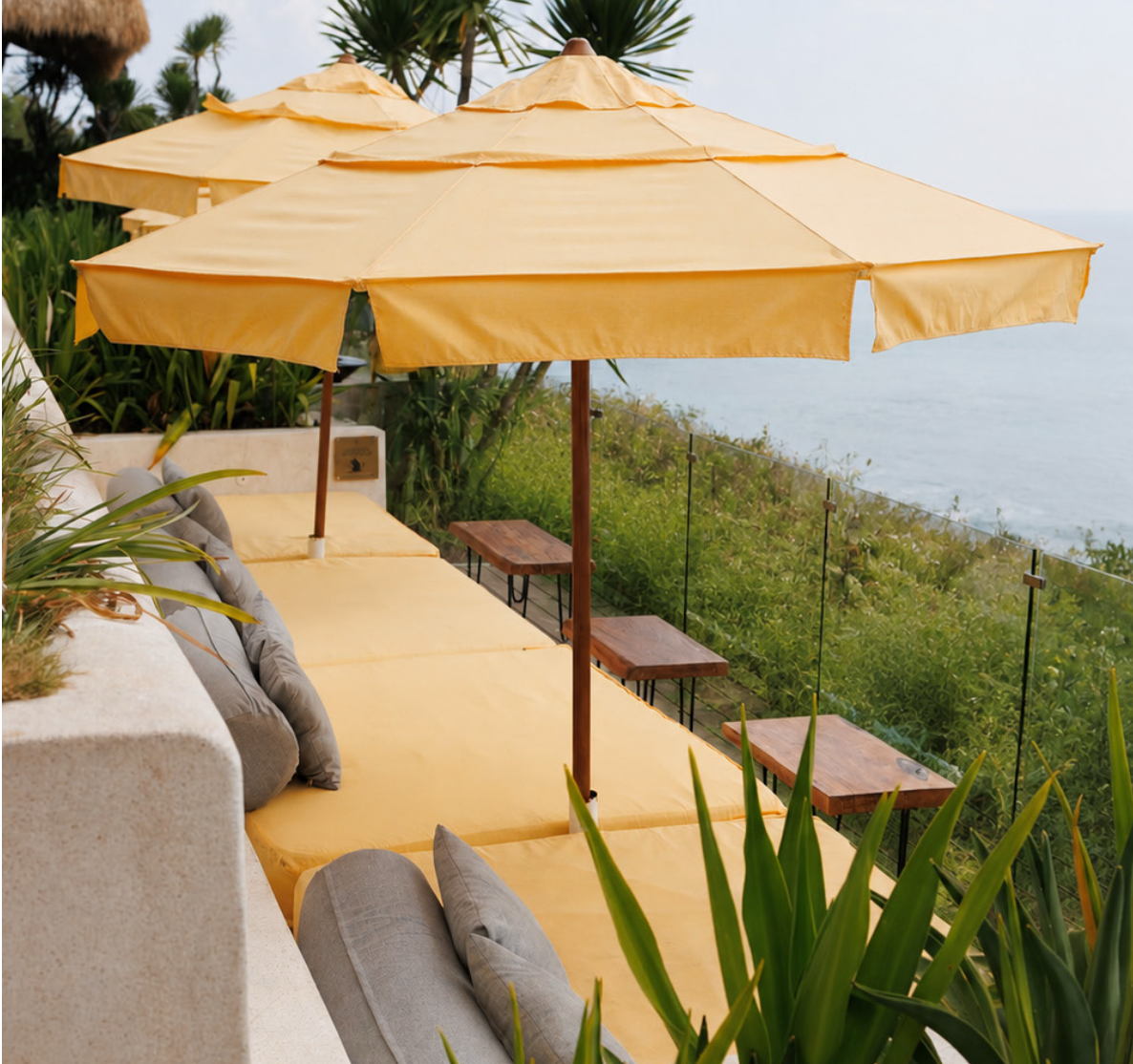 Outdoor lounge area with yellow umbrellas, cushioned seating, and small wooden tables overlooking the ocean and lush greenery.