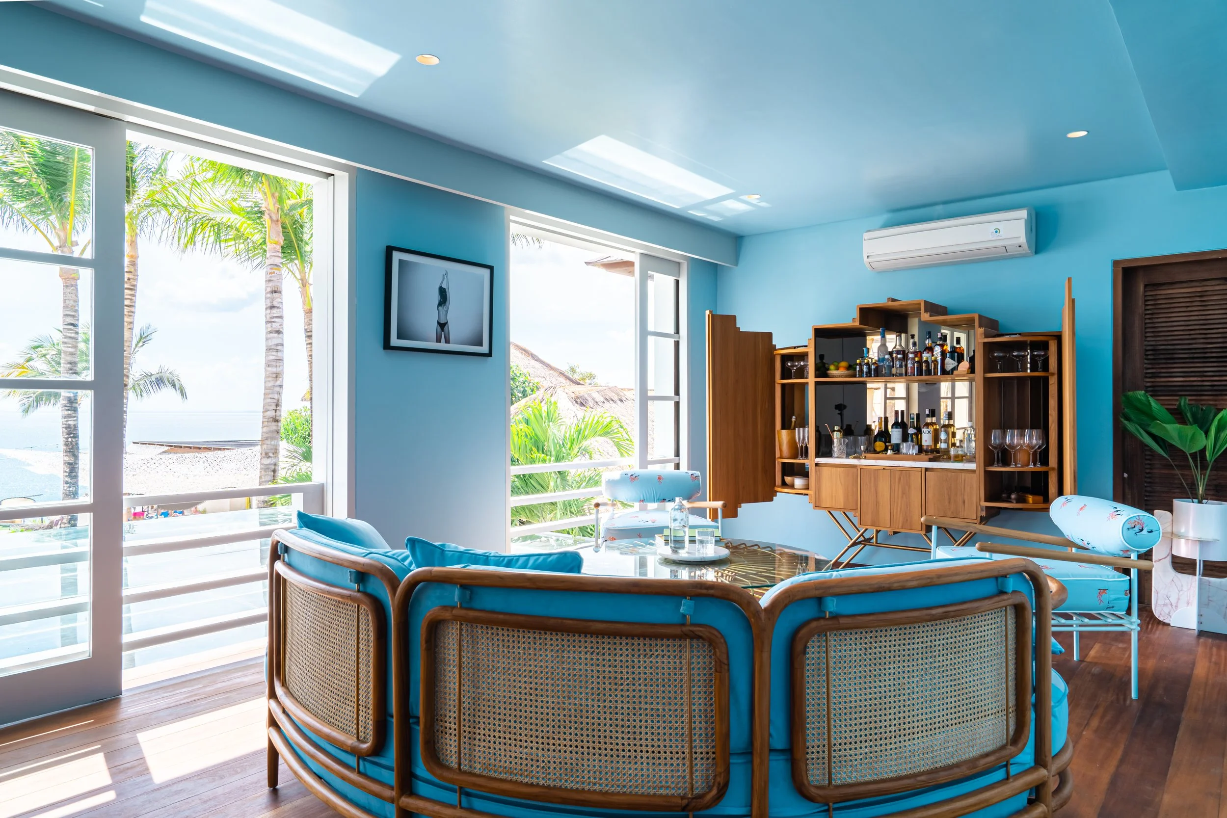 A bright, airy living room with large open windows and ocean view, featuring blue walls, wooden furniture, bar area, and tropical decor.