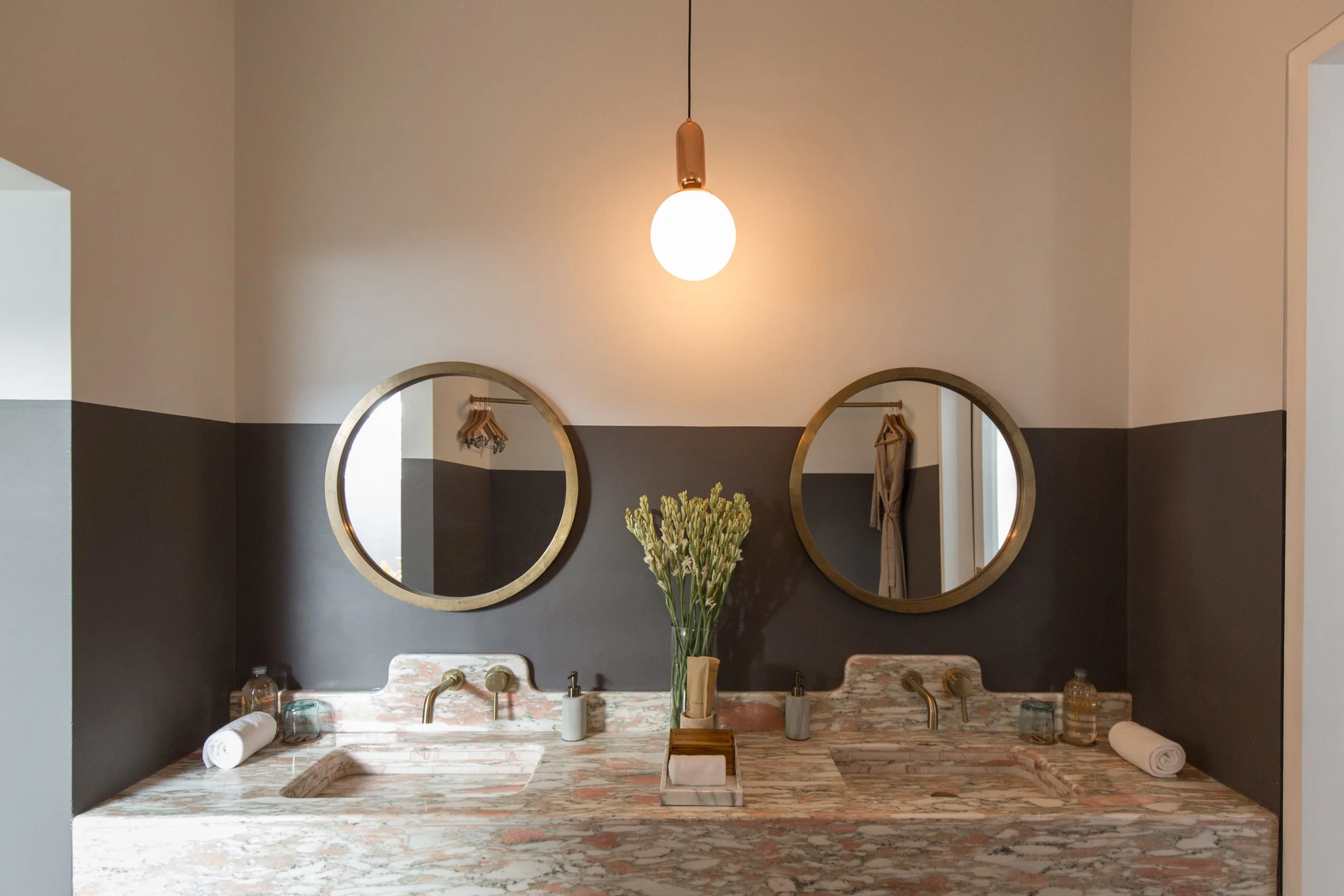 Bathroom with two round mirrors above a marble sink, a hanging light fixture, and a vase of flowers.