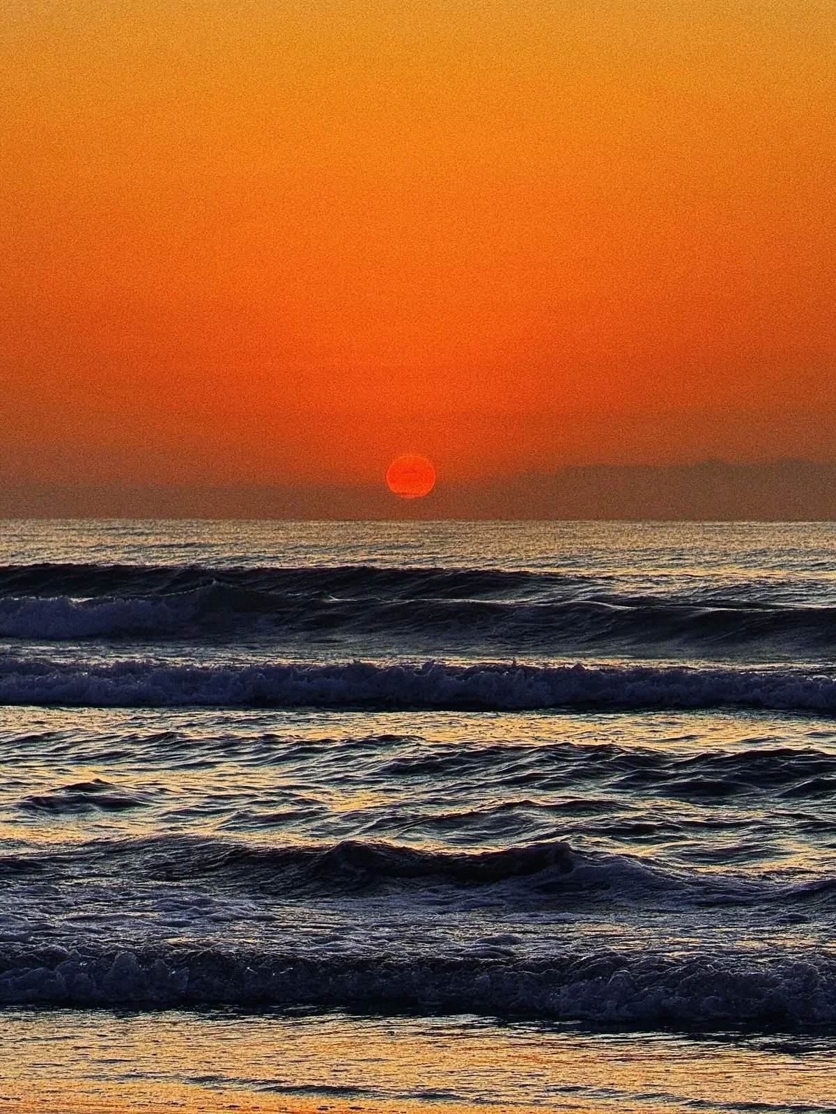 Sun setting over the ocean with waves in the foreground and a clear orange sky.