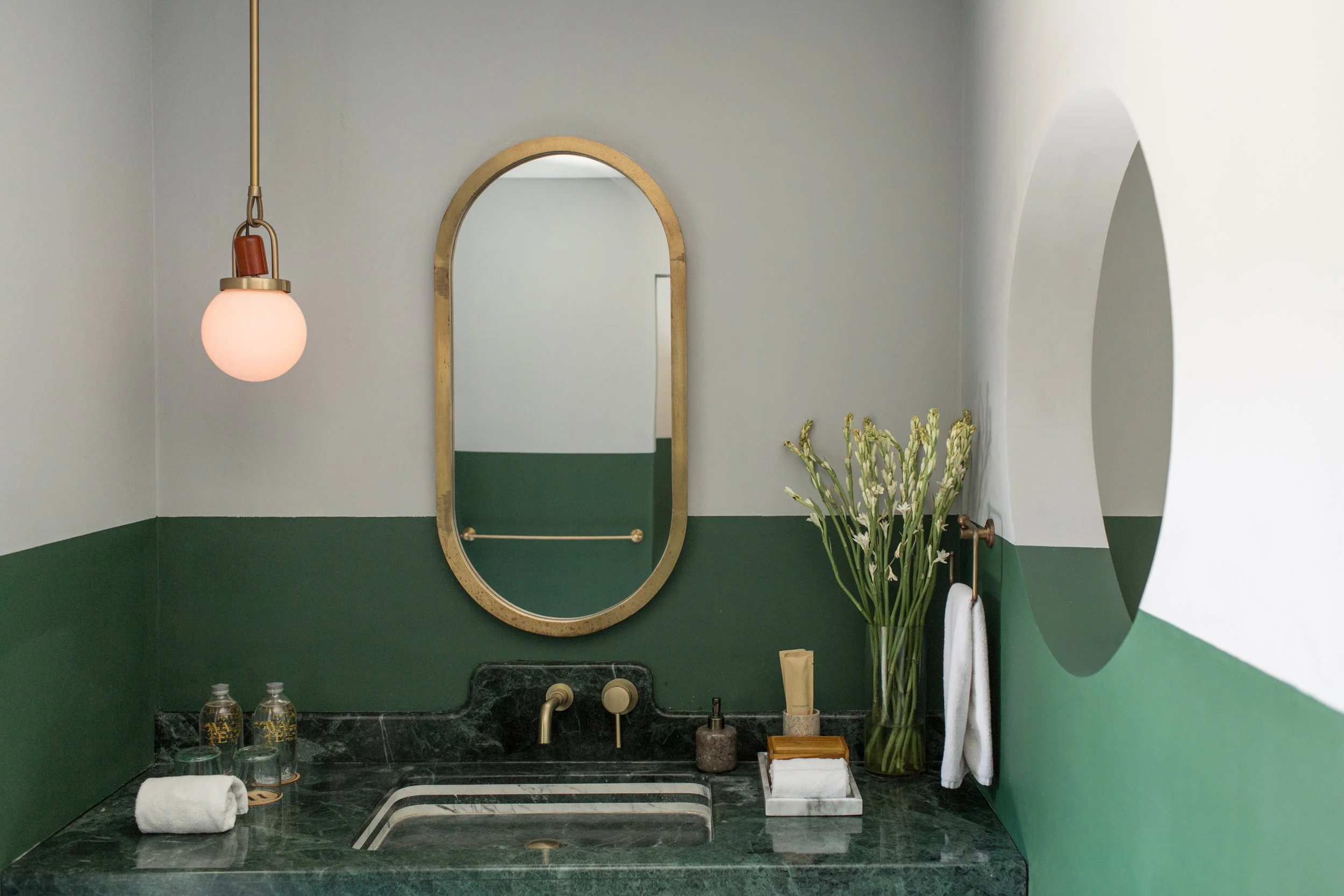 A bathroom sink area with a green and white color scheme, a gold-framed mirror, a hanging light fixture with a pink glass globe, a vase with white flowers, a bar of soap, and two water bottles on the sink counter.