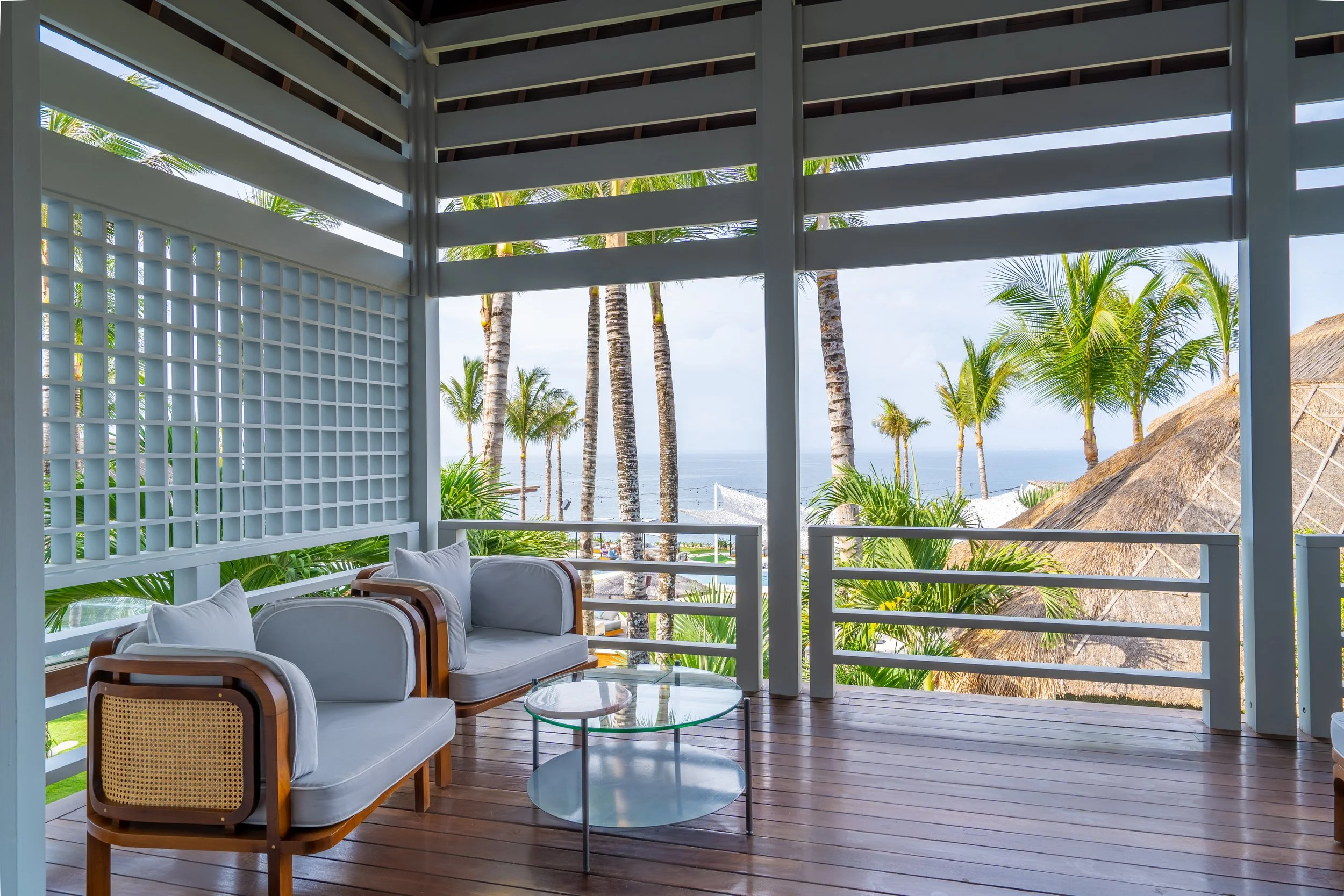 Outdoor lounge area with white cushioned chairs, glass coffee table, and a view of palm trees and the ocean through open fencing and thatched-roof structures.