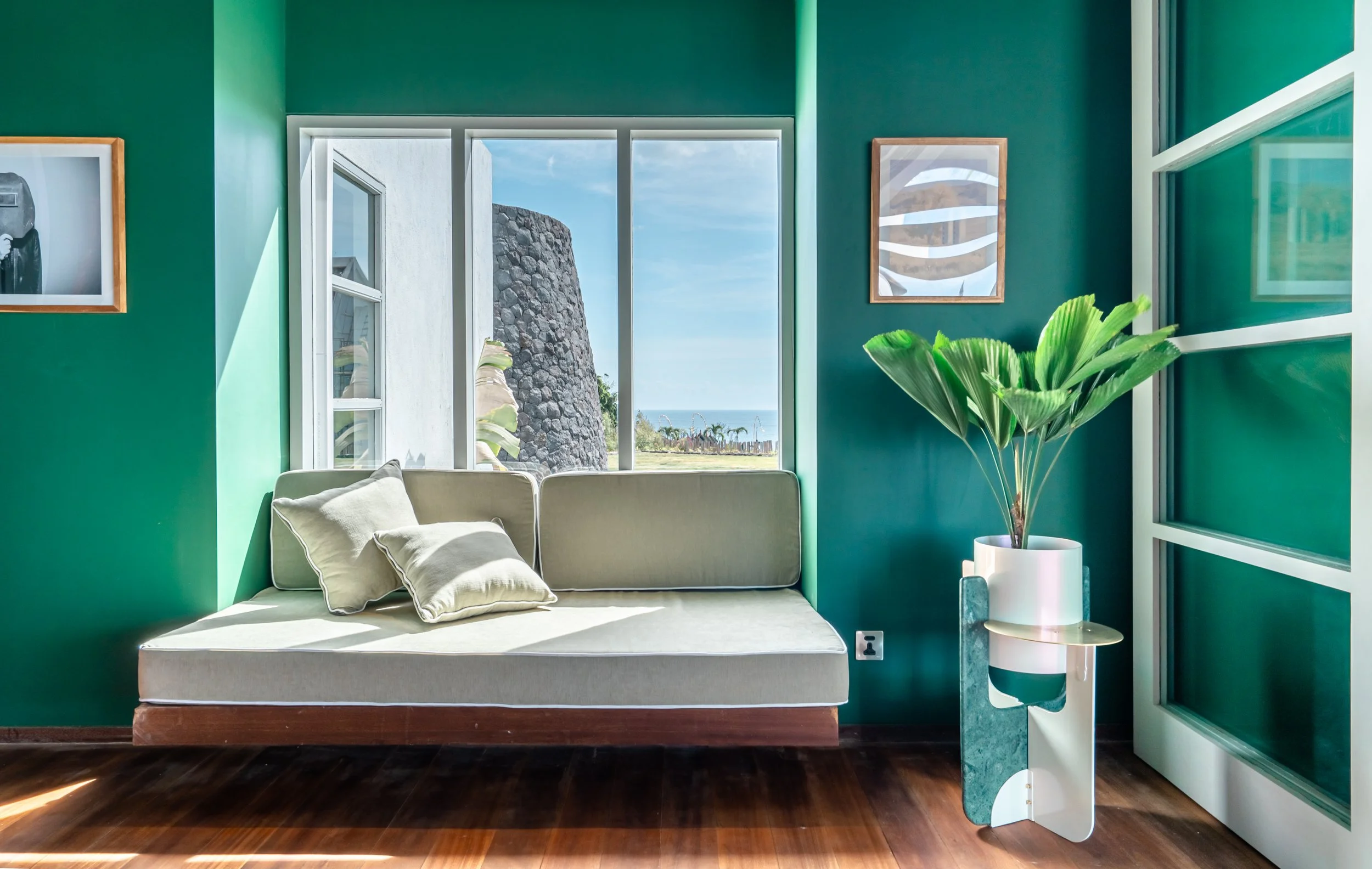 Interior of a room with teal walls, white window, and paintings. A beige sofa with matching pillows sits against the wall next to a large green plant in a white vase. Outside the window, a stone wall and a view of palm trees and the ocean can be seen