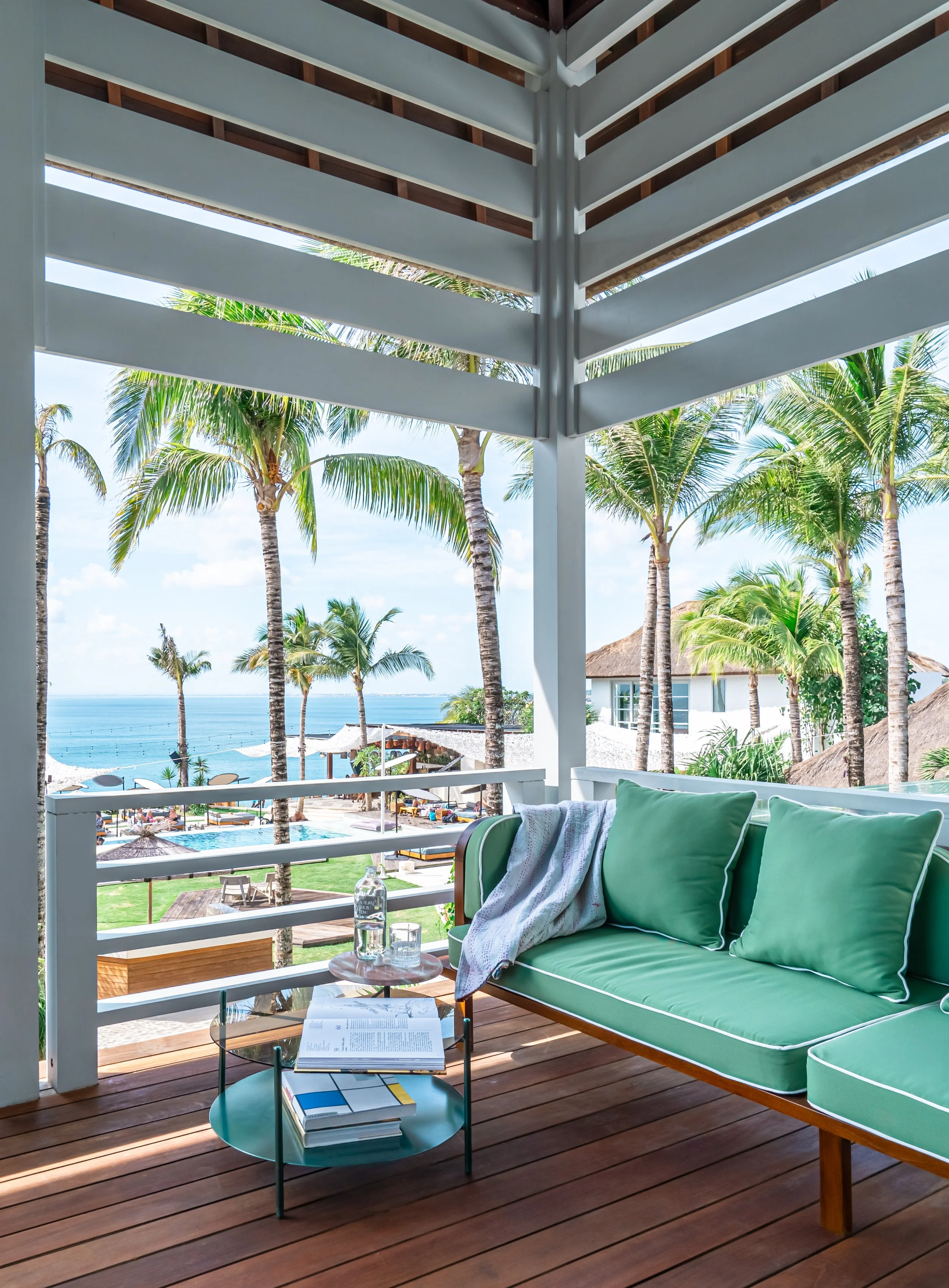 View of a balcony with a green cushioned sofa, a small round glass table with books and notebooks, and a bottle of water. In the background, tropical palm trees, a pool area with umbrellas and lounge chairs, and the ocean beyond.