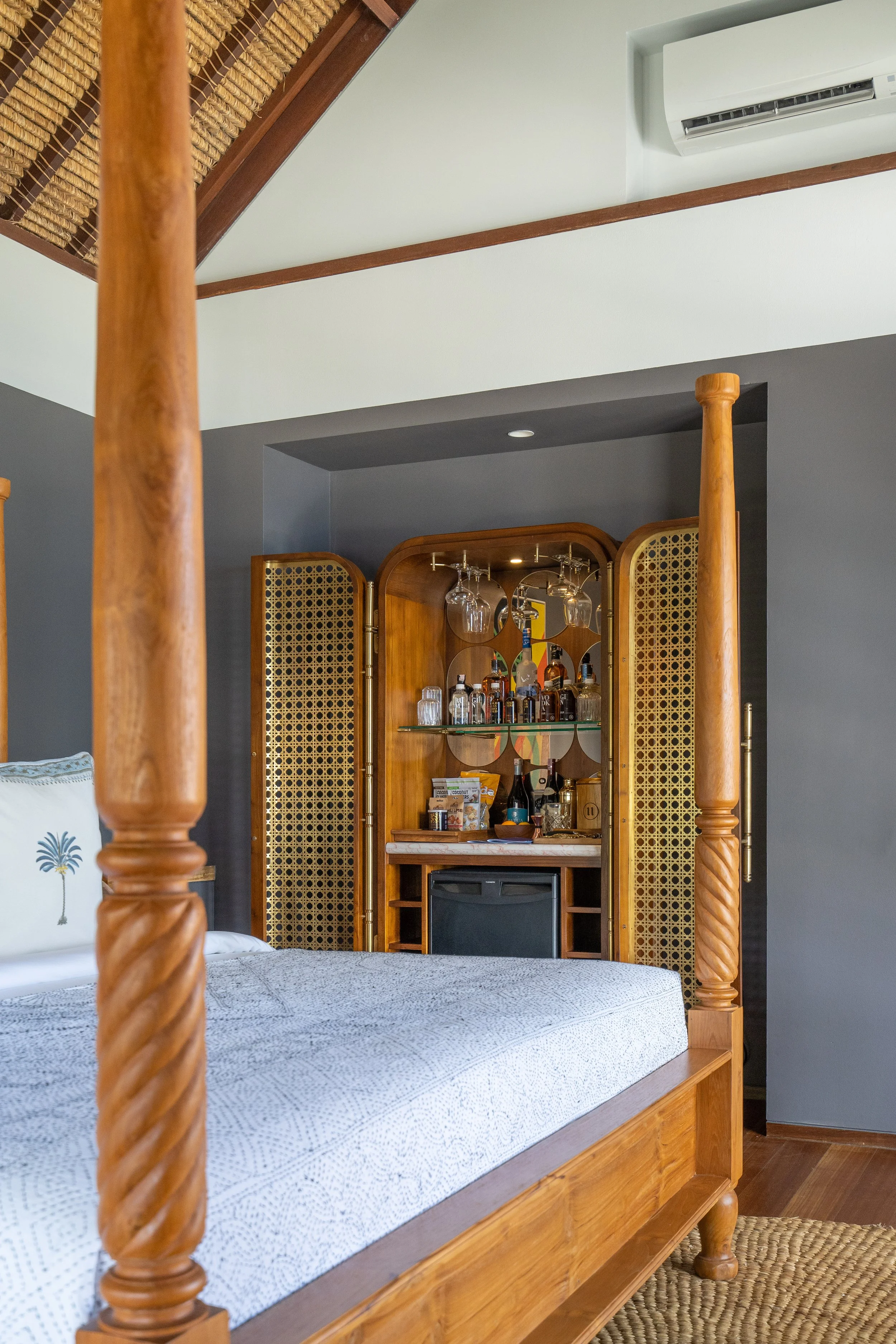 A bedroom with a wooden four-poster bed with a white textured bedspread, a beige braided rug, and a built-in wooden bar with glassware and bottles. There is an air conditioner mounted on the wall and a pillow with a pineapple embroidery on the bed.