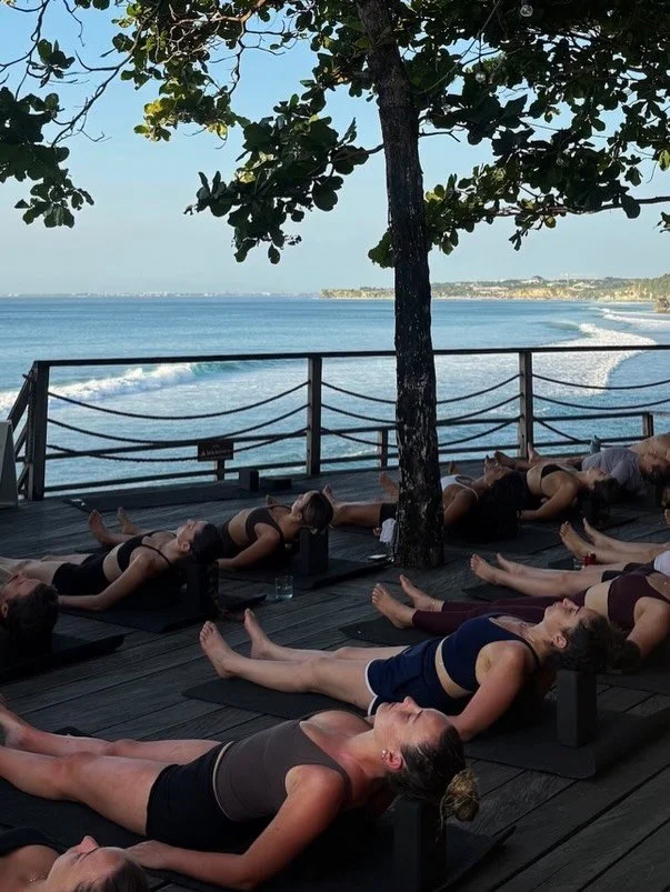 Group of women practicing yoga on a wooden deck by the ocean, with a tree in the center and a view of the water and coastline in the background.