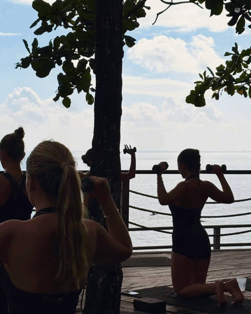 Women practicing yoga on a wooden deck by the water with a tree in the foreground and a cloudy sky in the background.