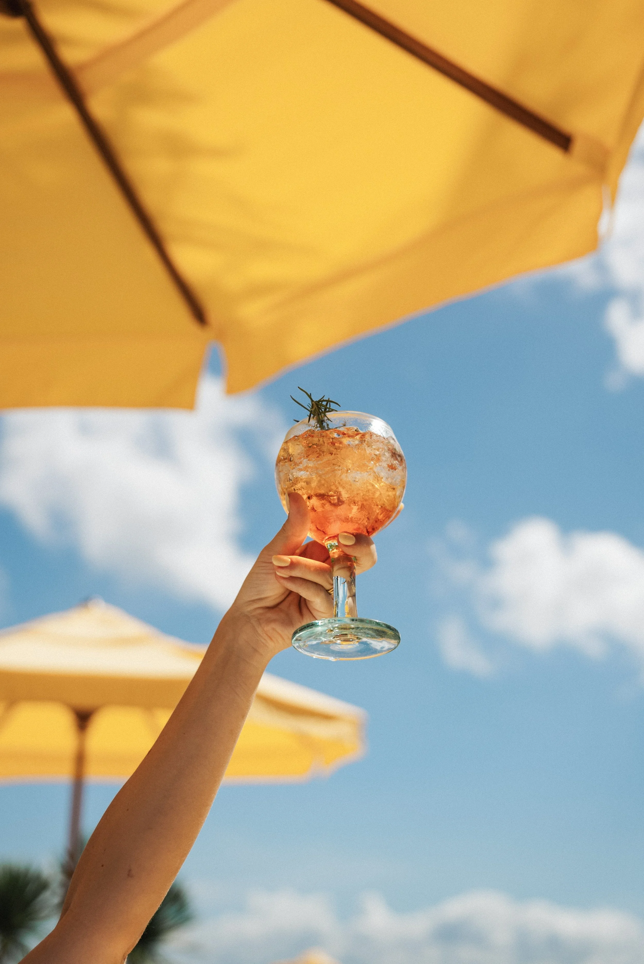A hand holding a glass of pink-colored cocktail with ice and a sprig of rosemary under a yellow umbrella on a bright sunny day at the beach.