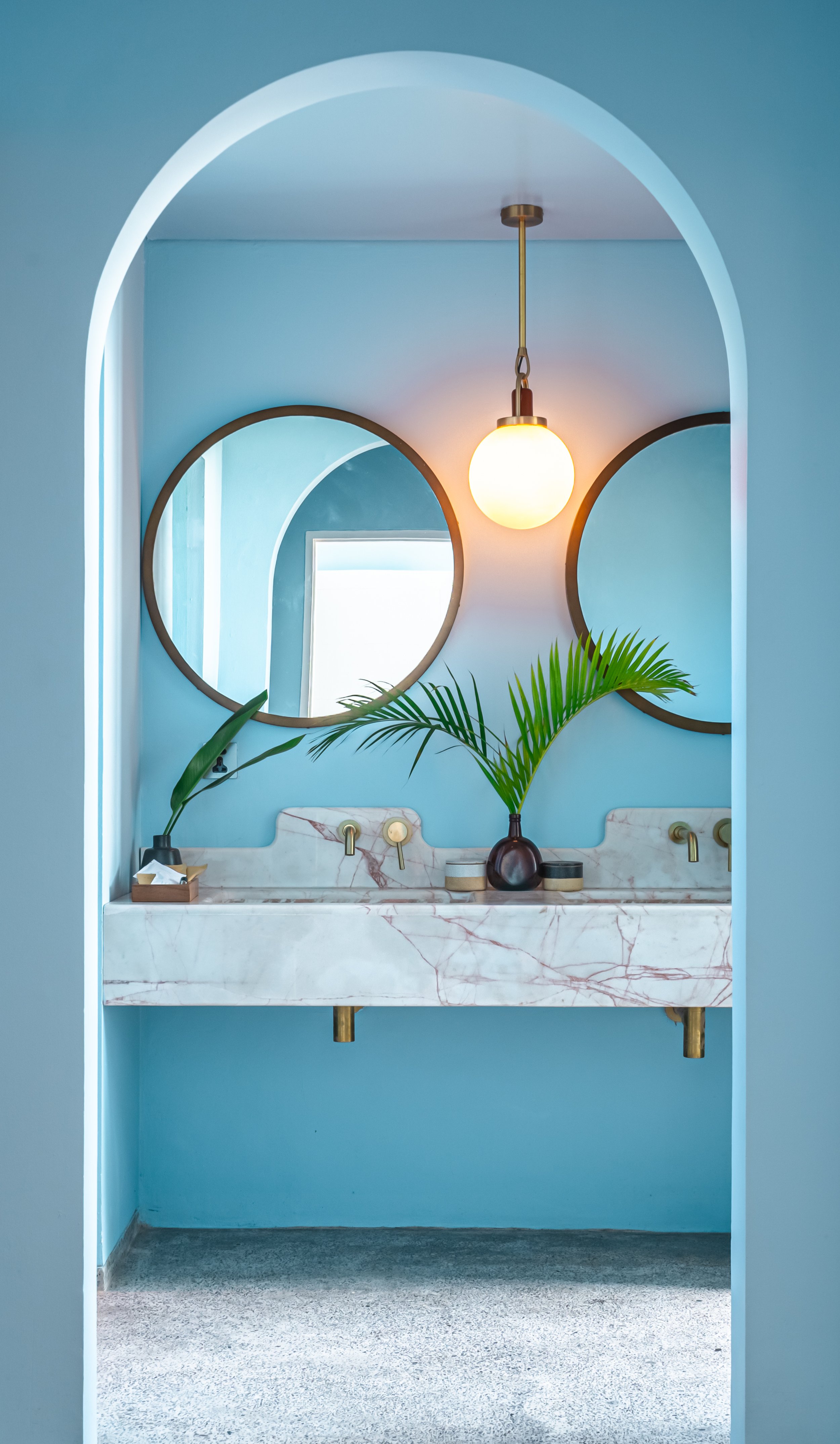 Bathroom with blue walls, featuring a white marble sink with gold fixtures, a large glowing pendant light, two circular mirrors, and green plants in dark vases.
