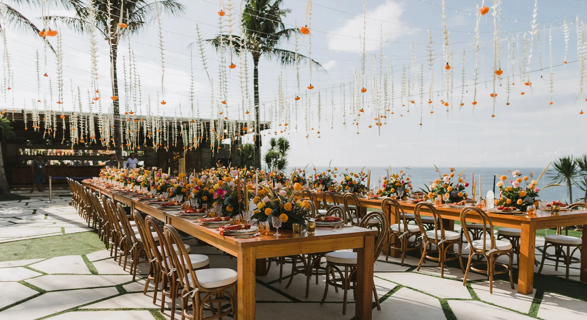 Decorated outdoor event space with long banquet table featuring floral centerpieces, hanging orange and white flowers, and view of the ocean in the background.
