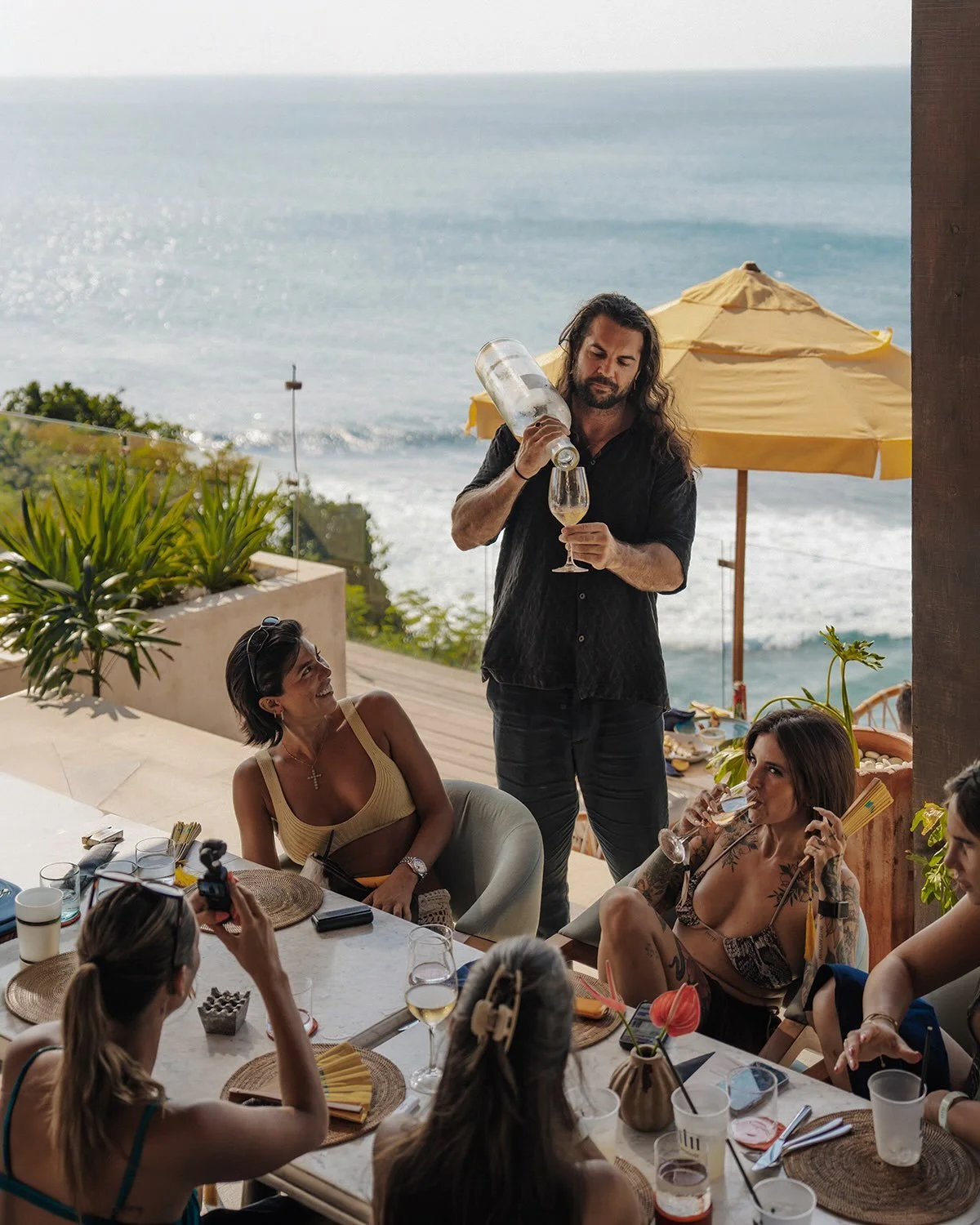 A man with long hair pouring wine into a glass for a woman sitting at an outdoor dining table with friends, with a beach and ocean in the background.