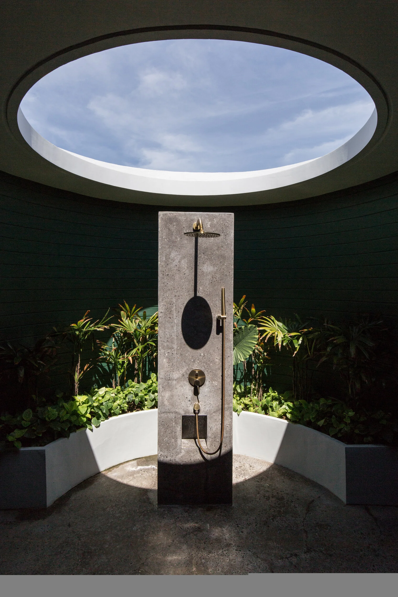 An outdoor shower with a circular roof opening showing a blue sky with clouds, surrounded by green plants and a white curved wall.