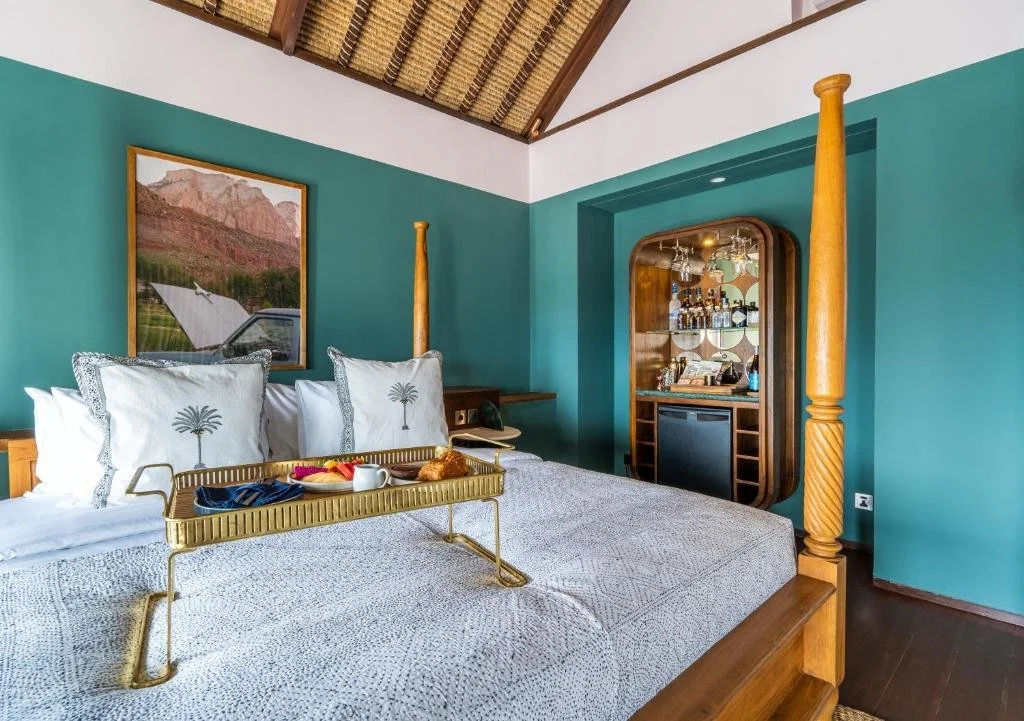 A bedroom with teal walls, a wooden four-poster bed with white and patterned pillows, a tray with breakfast on the bed, and a built-in bar area with bottles and glasses.