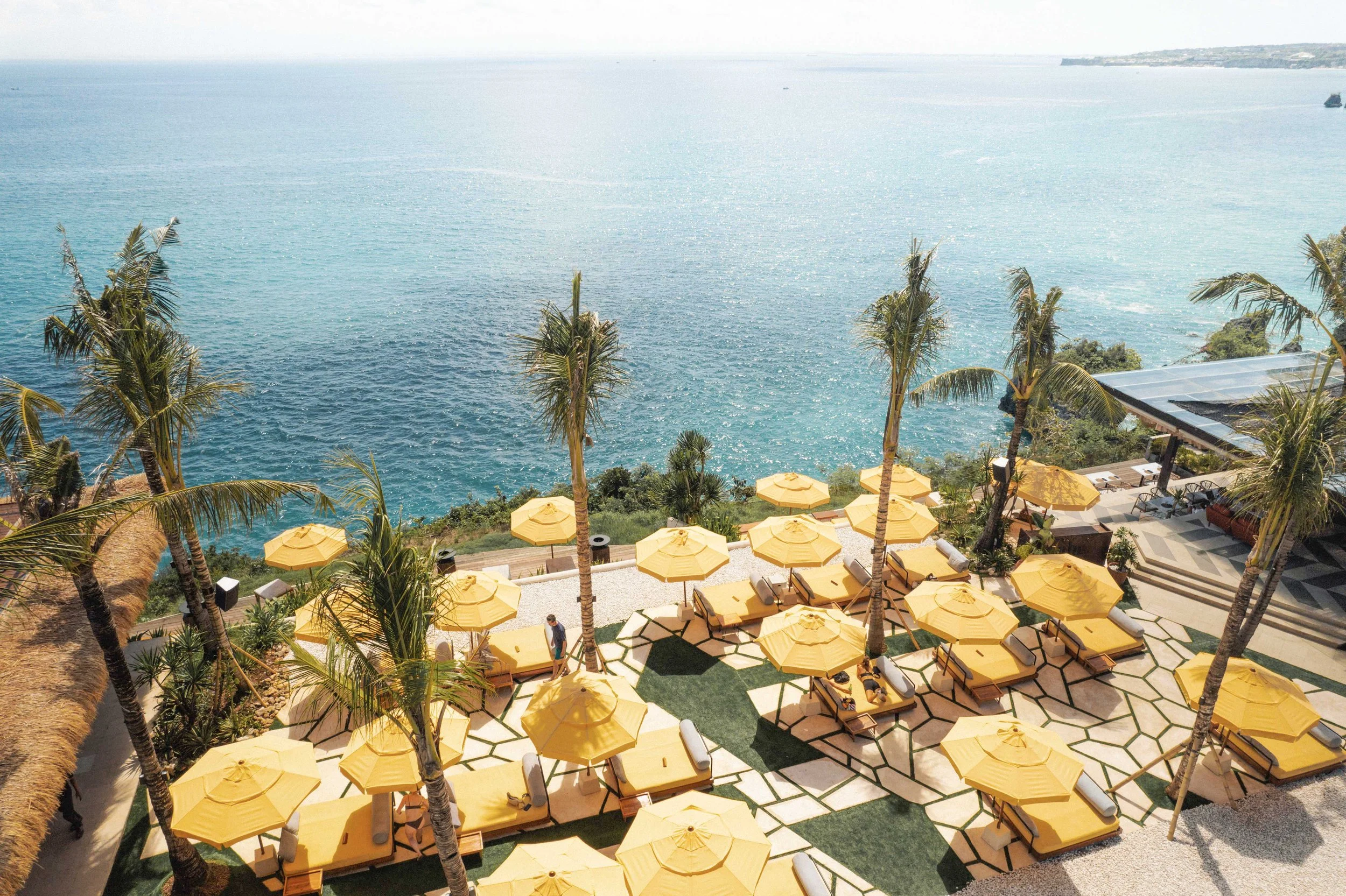 A scenic view of a seaside resort with yellow umbrellas and lounge chairs on a patio, palm trees, and the ocean in the background.