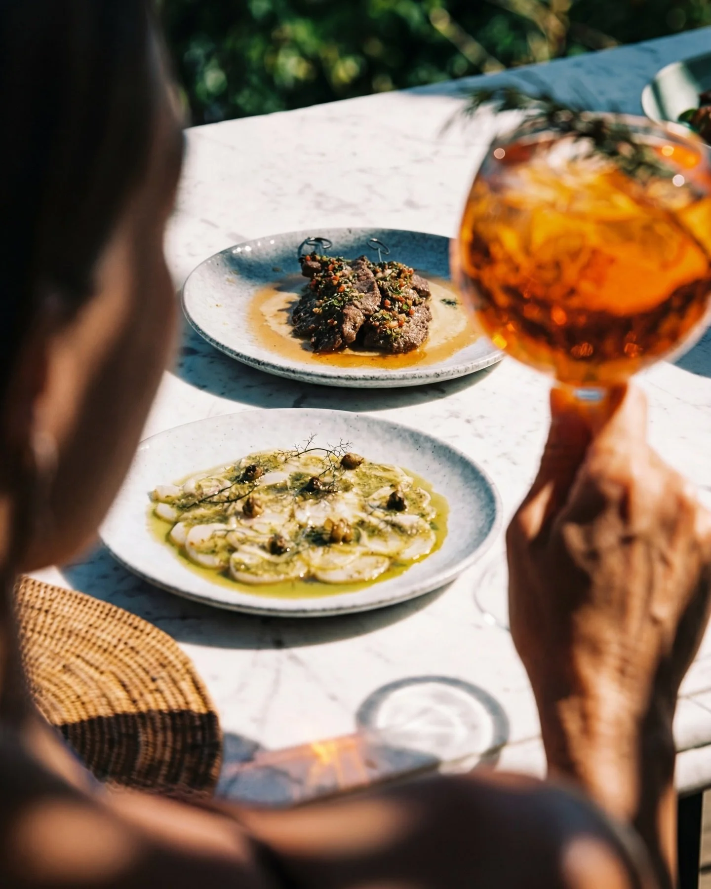 A woman holding a glass of what appears to be rosé wine, with three plates of food on a white table outdoors. The food includes a dish with sliced vegetables, a cooked meat dish, and another with what looks like seafood or fish, garnished with herbs.