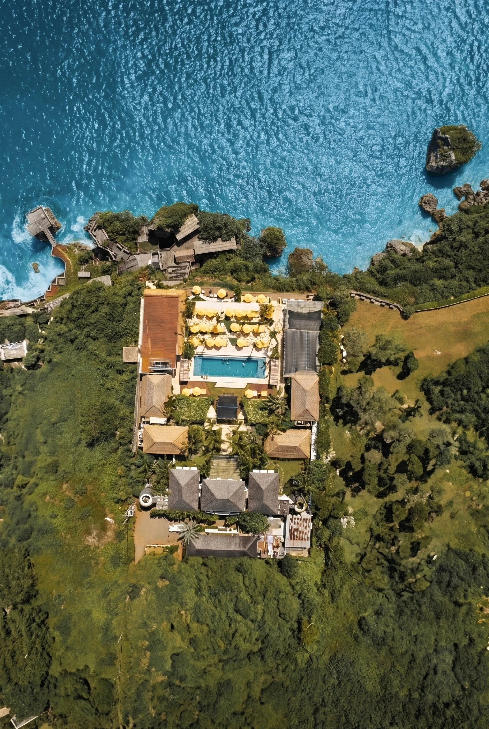 An aerial view of a coastal area showing a large blue ocean with a small rocks and greenery, and a coastline with resorts, gardens, and buildings.