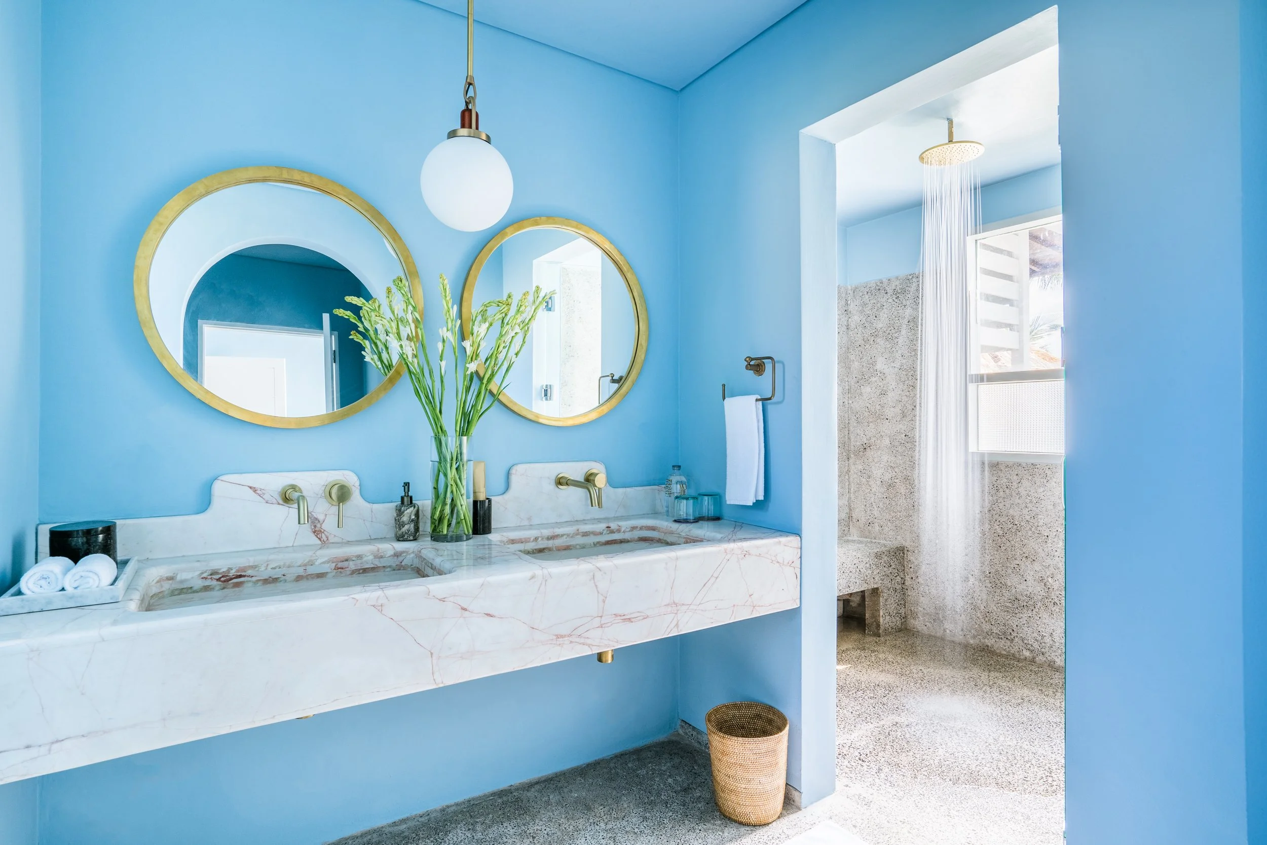 A modern bathroom with light blue walls, a marble vanity with gold fixtures, and two round mirrors with gold frames. A vase with green flowers sits on the vanity. There is a white towel hanging on a gold towel rack and a wicker basket on the floor. T
