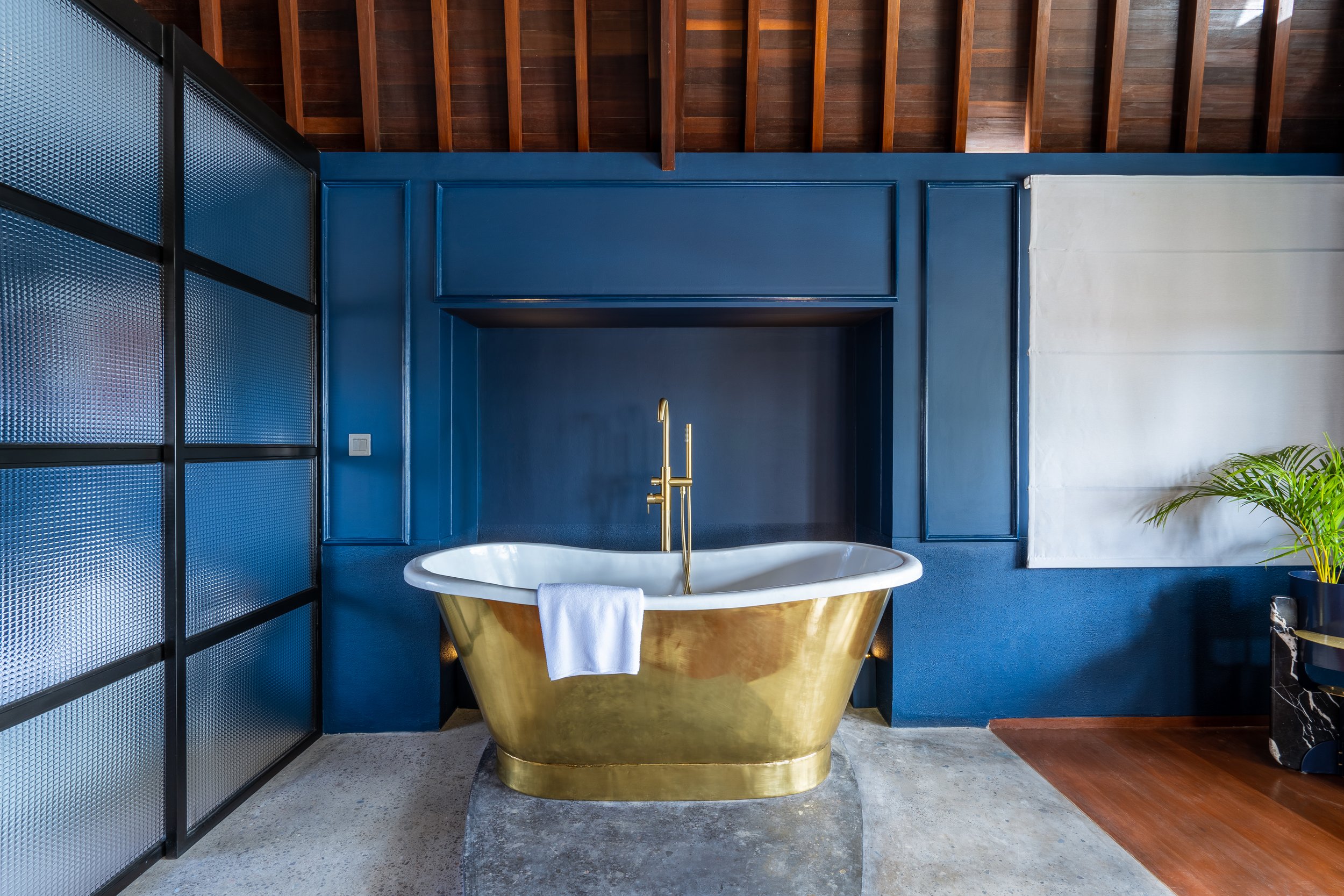 A luxurious bathroom with a gold bathtub, brass faucet, and a white towel hanging over the tub. The room features a deep blue wall with decorative paneling, a textured glass partition, exposed wooden ceiling beams, and a potted plant in the corner.