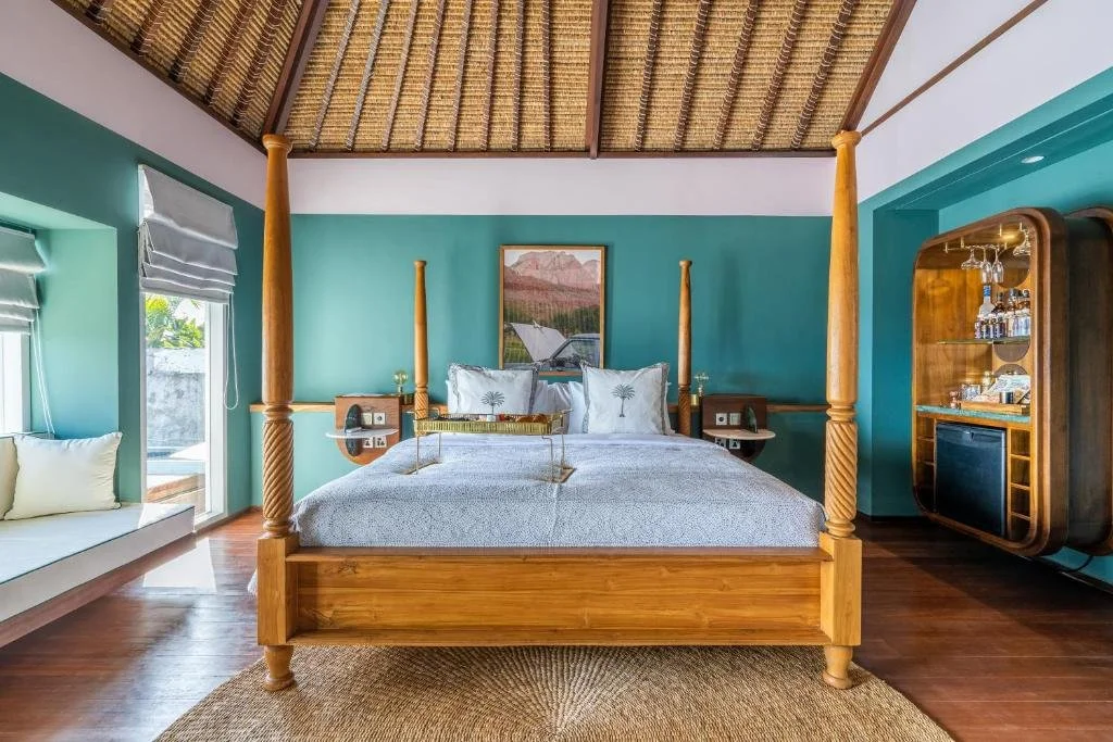 A bedroom with a four-poster wooden bed, teal walls, a picture above the bed, windows with blinds, a built-in window seat with pillows, a bar cabinet with glassware, and wooden flooring with a rug.