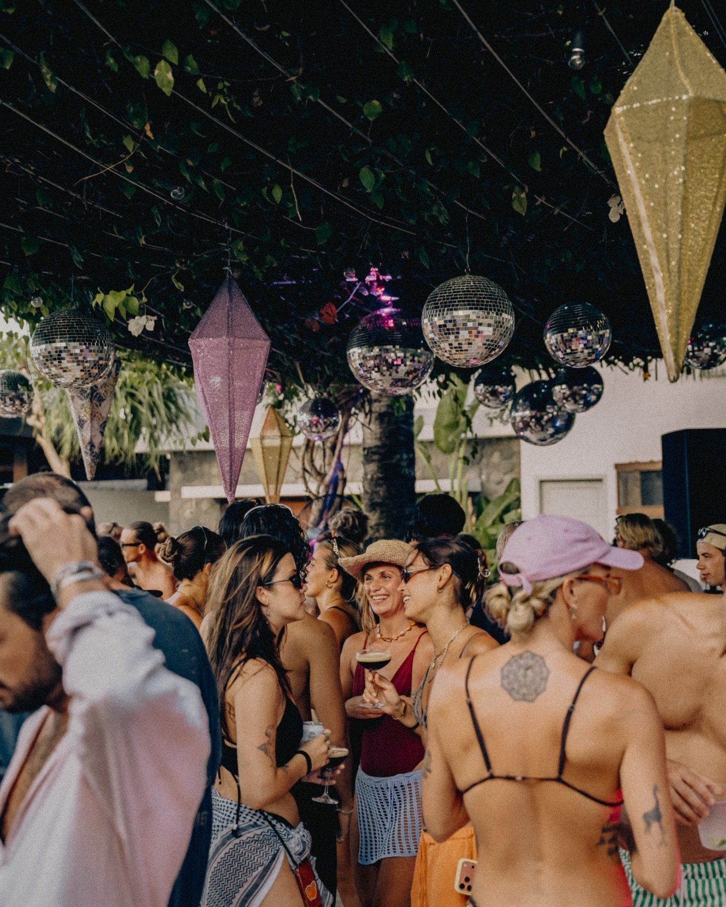 A crowded outdoor party scene with people socializing under a canopy decorated with hanging disco balls and colorful paper lanterns. People are dressed in summer attire, some holding drinks, and the atmosphere appears lively and festive.
