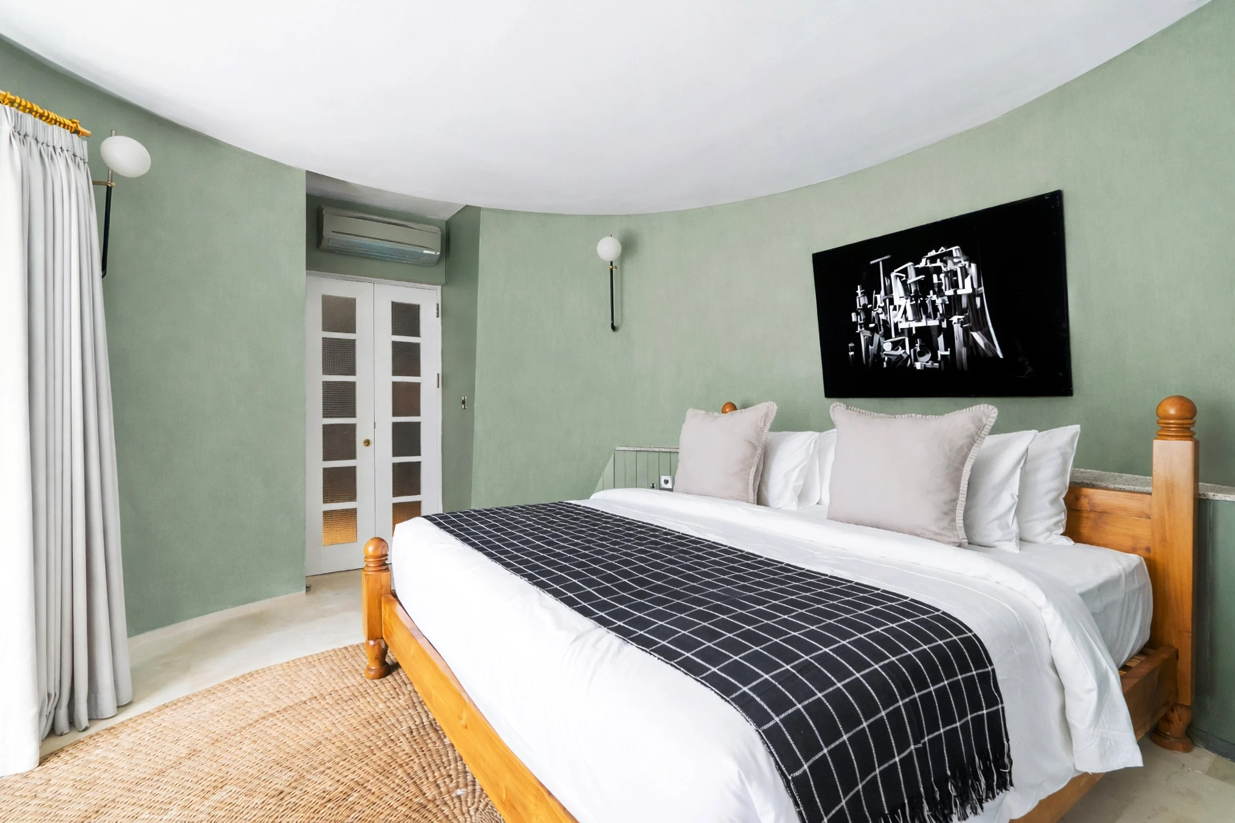 A bedroom with green walls, a wooden bed with white and beige pillows, and a black-and-white checkered blanket. There is a black wall-mounted art piece, white curtains, a door, and an air conditioning unit above the door.