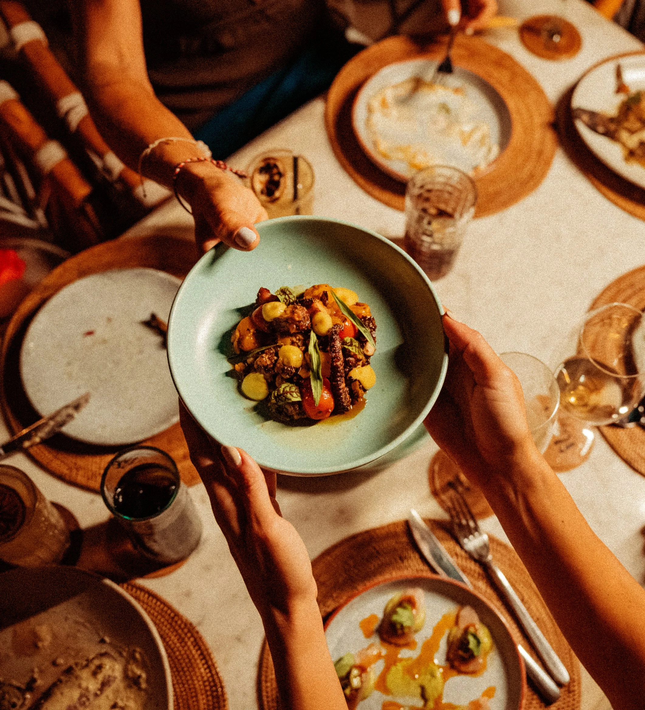 People holding a bowl of Mexican food, with various dishes and drinks on a table, in a cozy restaurant setting.