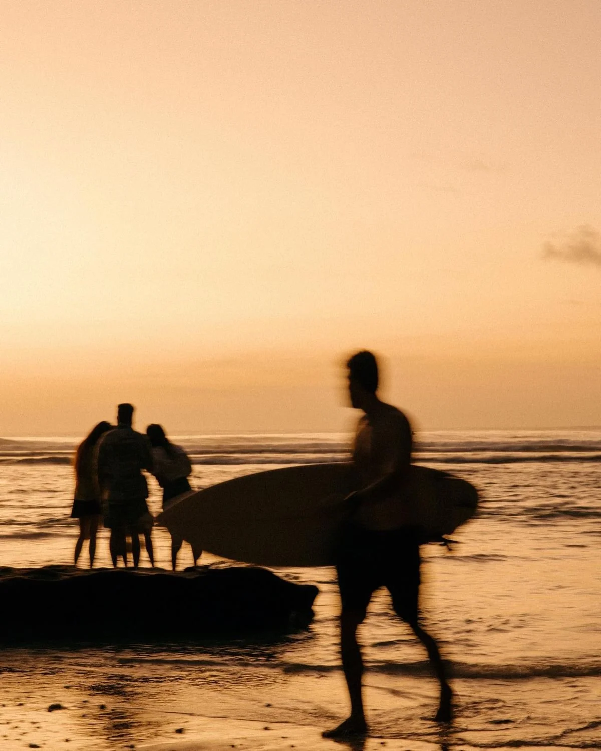 Silhouettes of people on the beach at sunset, with one person holding a surfboard and others near the water.