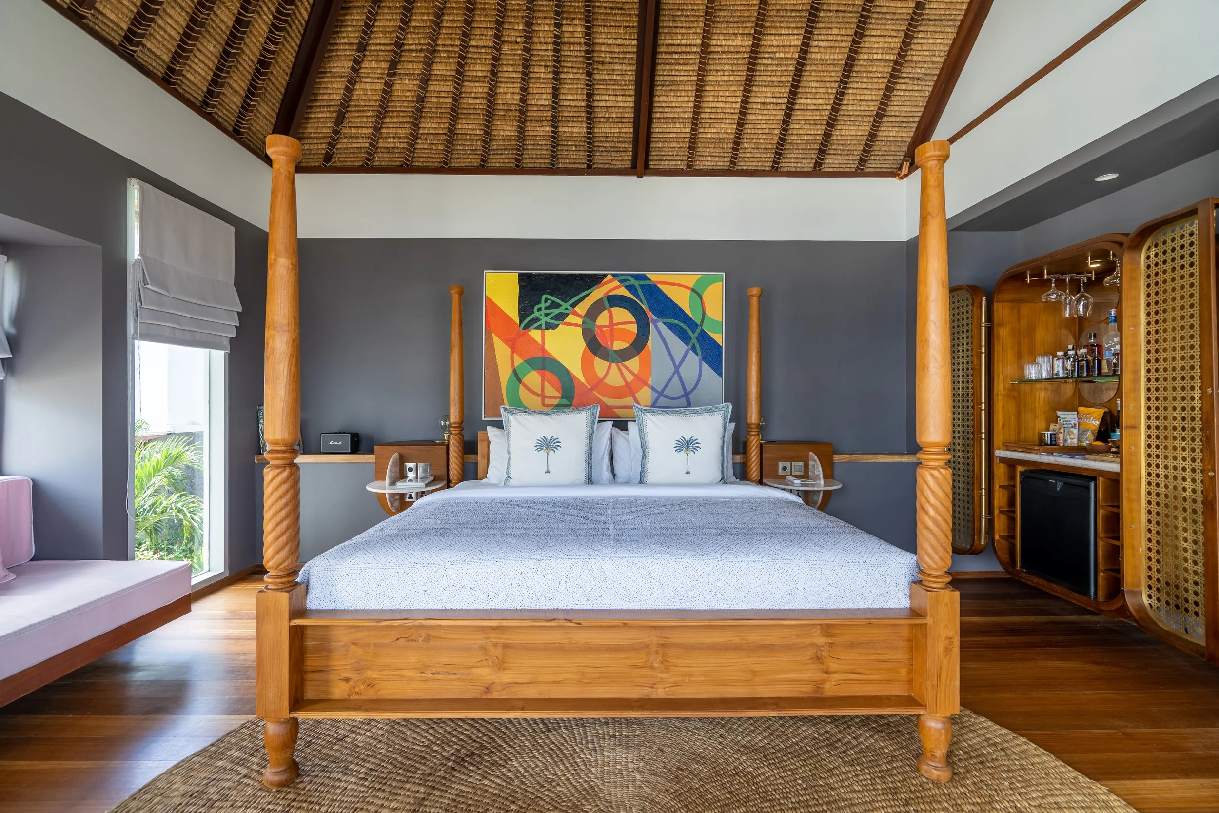 A bedroom with a four-poster wooden bed, grey walls, colorful abstract artwork above the bed, large windows with white blinds, a side table with electrical outlets, and a bar cabinet with wine glasses and bottles.