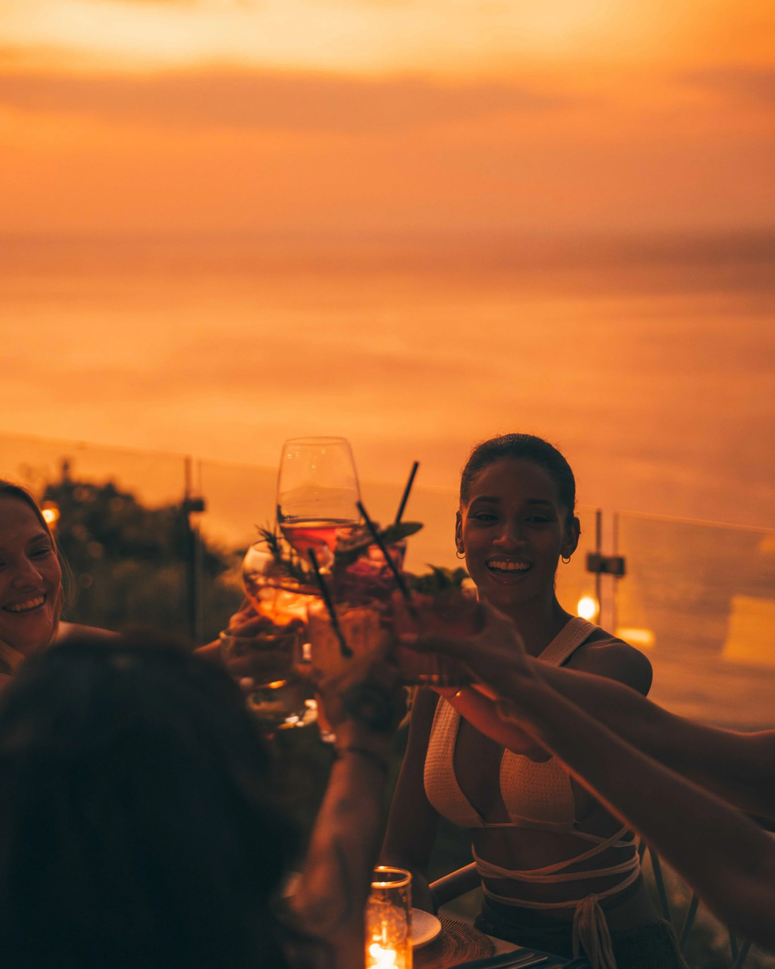People enjoying drinks at a sunset outdoor gathering, with a woman smiling and holding a glass of a cocktail, in a warm, orange-hued sky background.