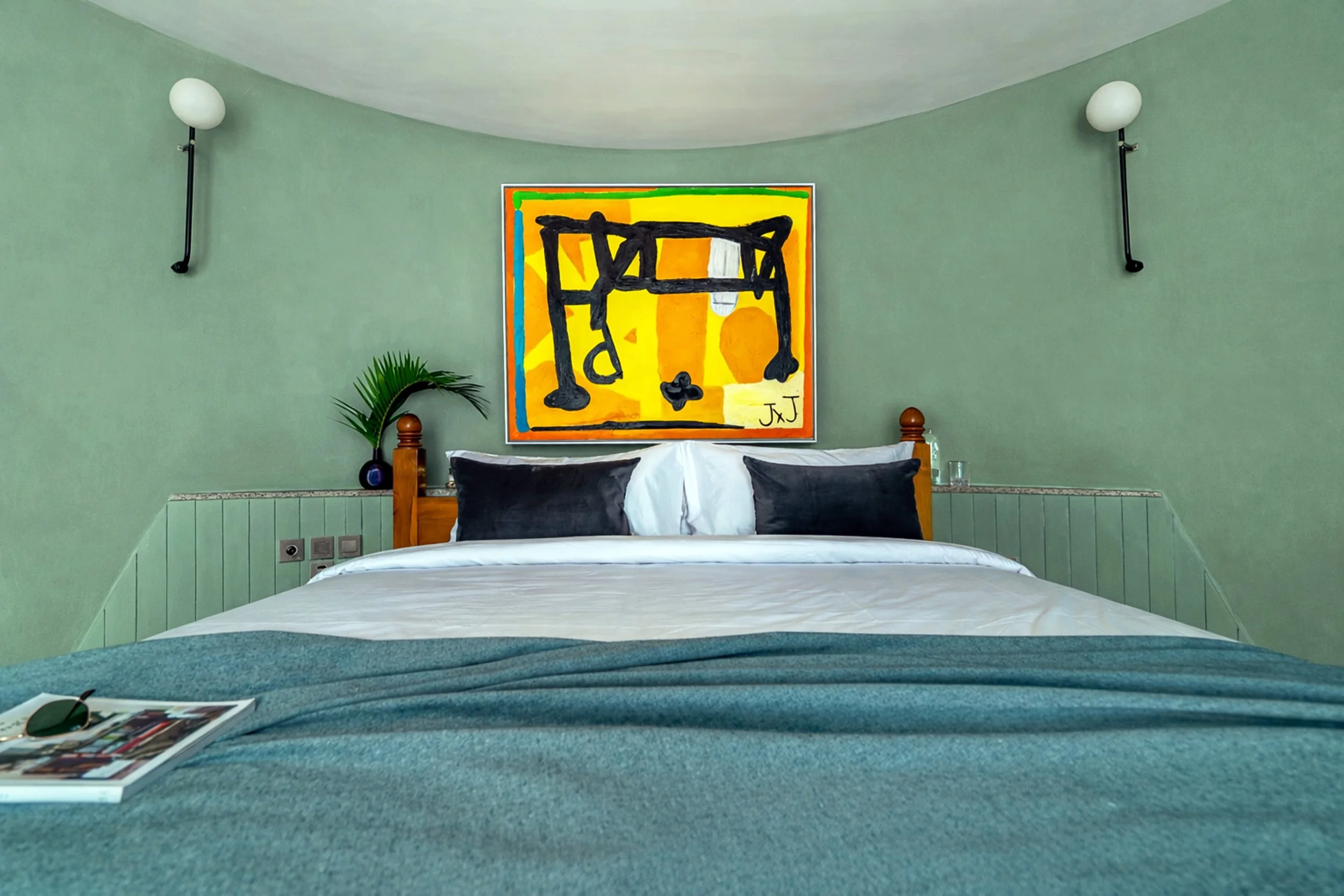 A hotel room with a bed, a colorful abstract painting above the headboard, two black and white pillows, a green plant, and wall-mounted lights on green walls.