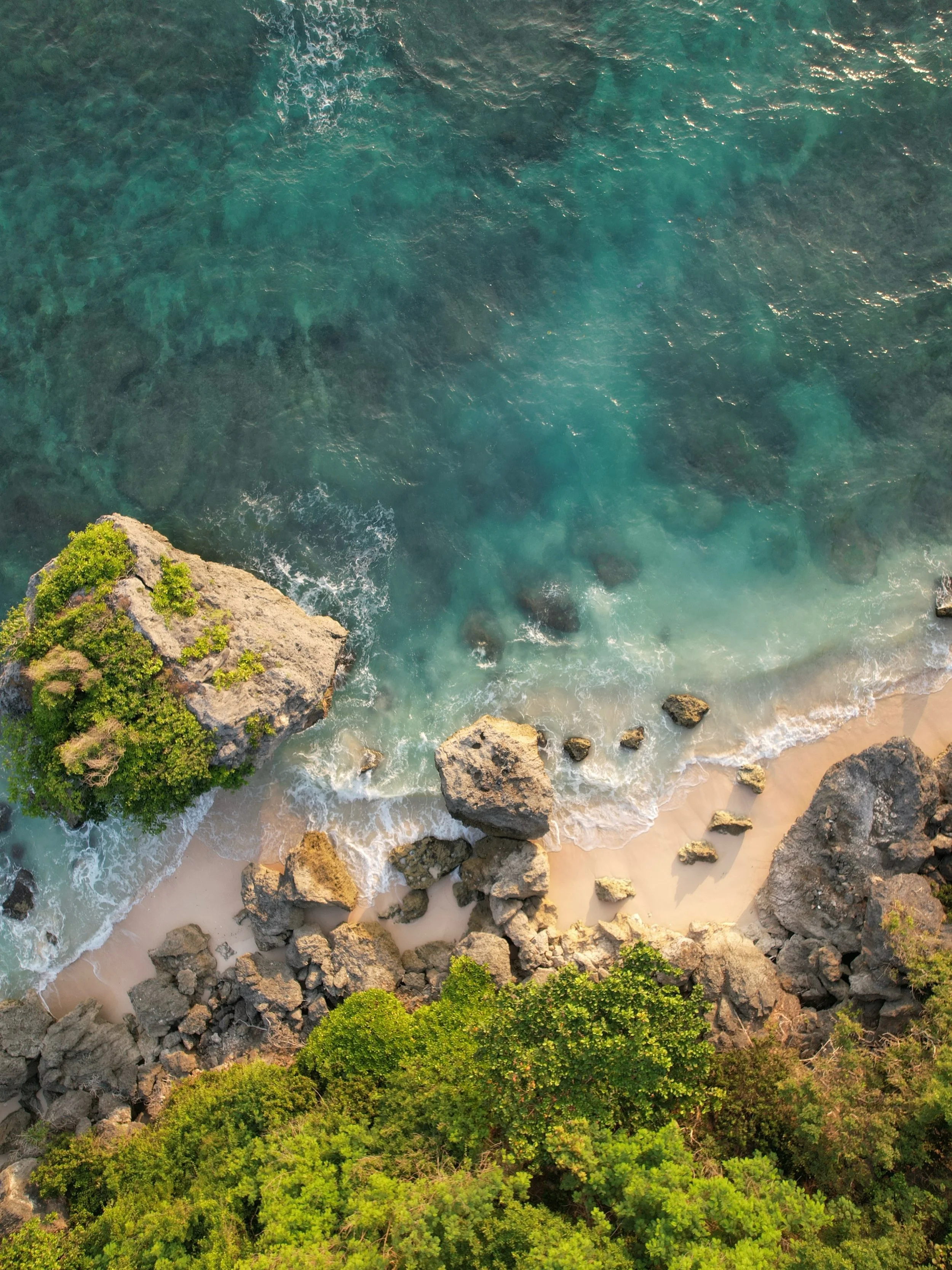 An aerial view of a rocky coastline with clear turquoise water, large rocks on the sandy shore, and green trees at the bottom of the image.