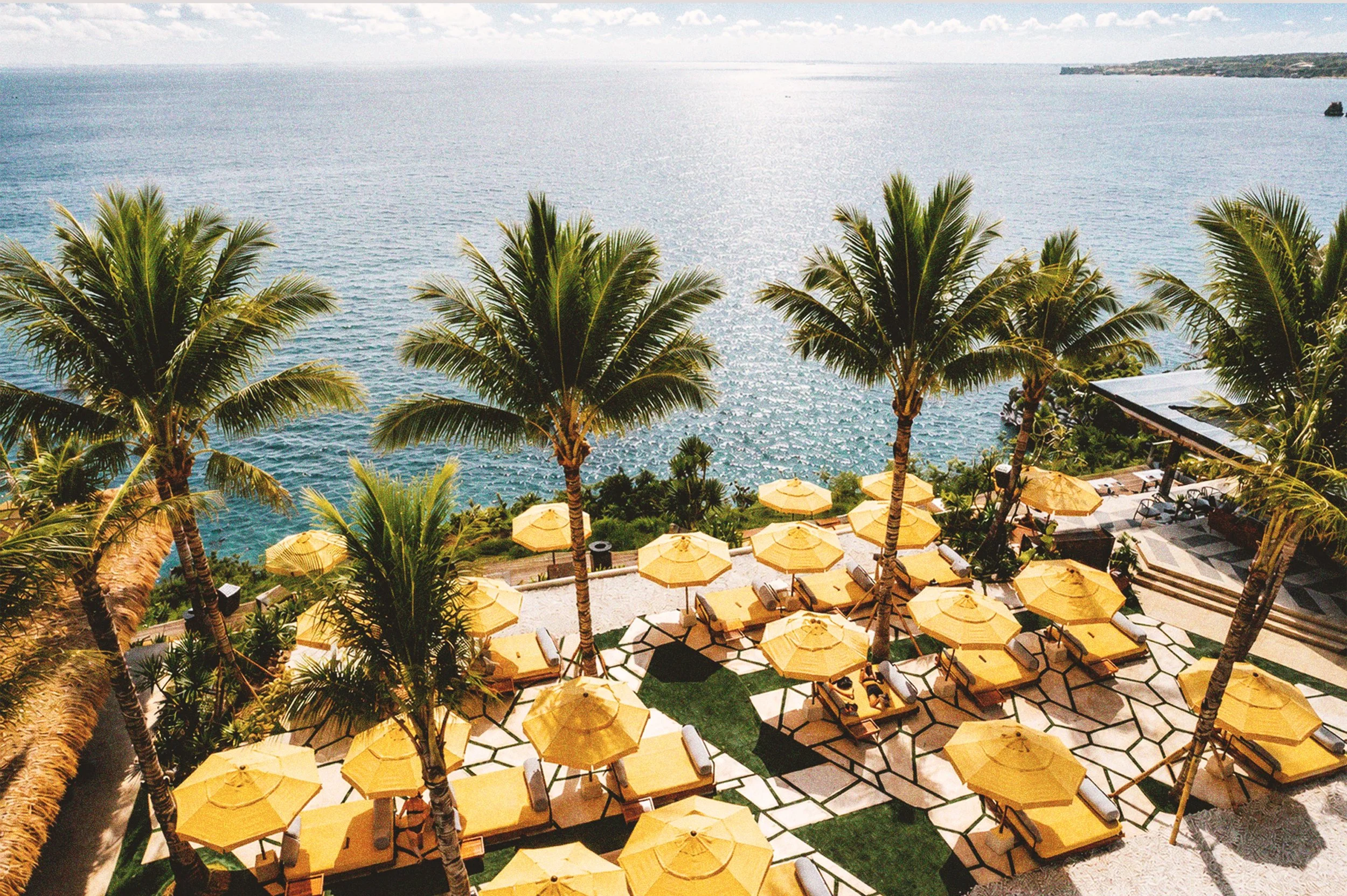 View of a seaside resort with palm trees, yellow umbrellas, and lounge chairs on a patio, overlooking the ocean with a distant ship and coastline.
