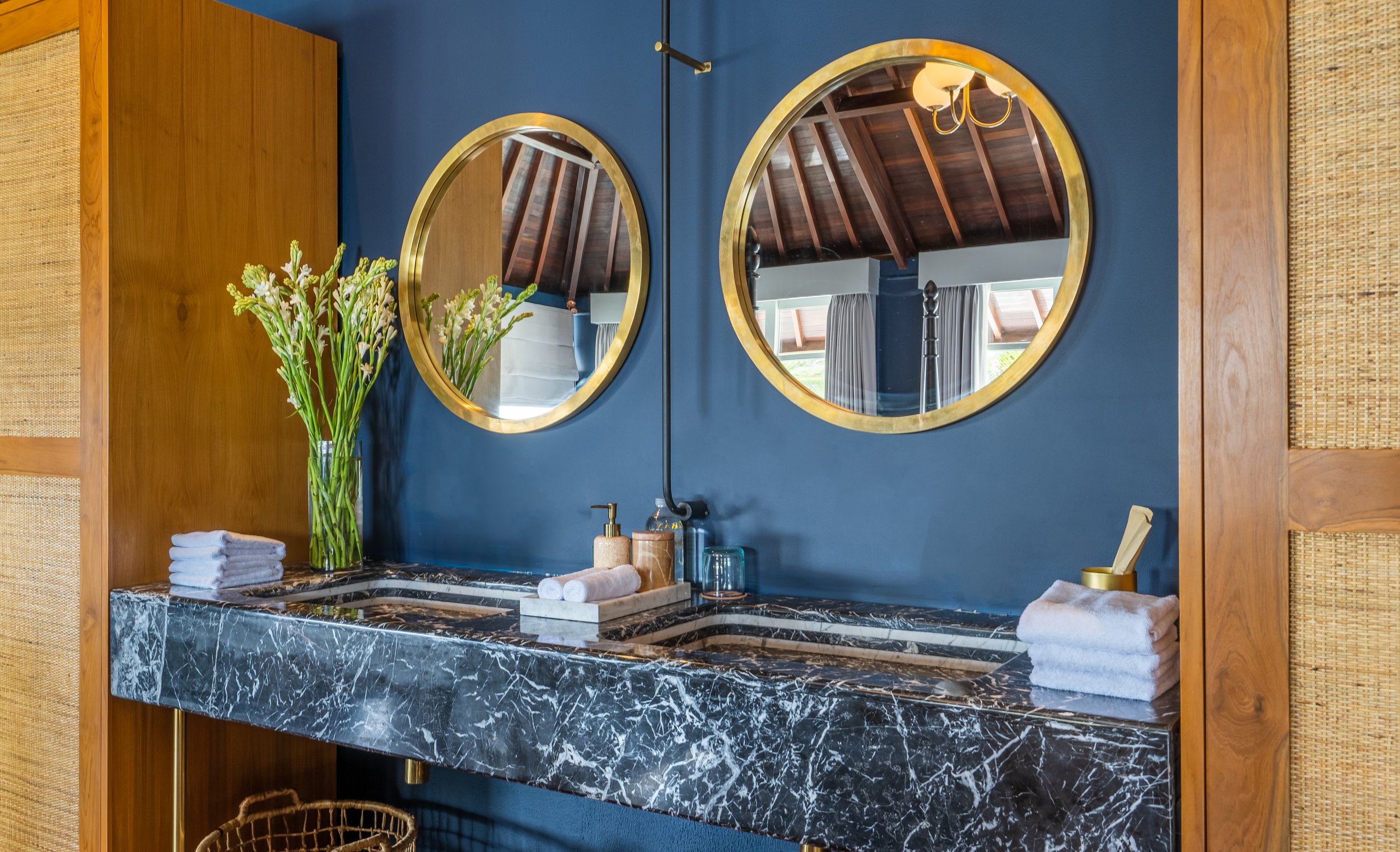 A bathroom vanity with a dark green marble countertop, two round gold-framed mirrors, a vase of white flowers, neatly folded towels, and toiletries, against a dark blue wall.
