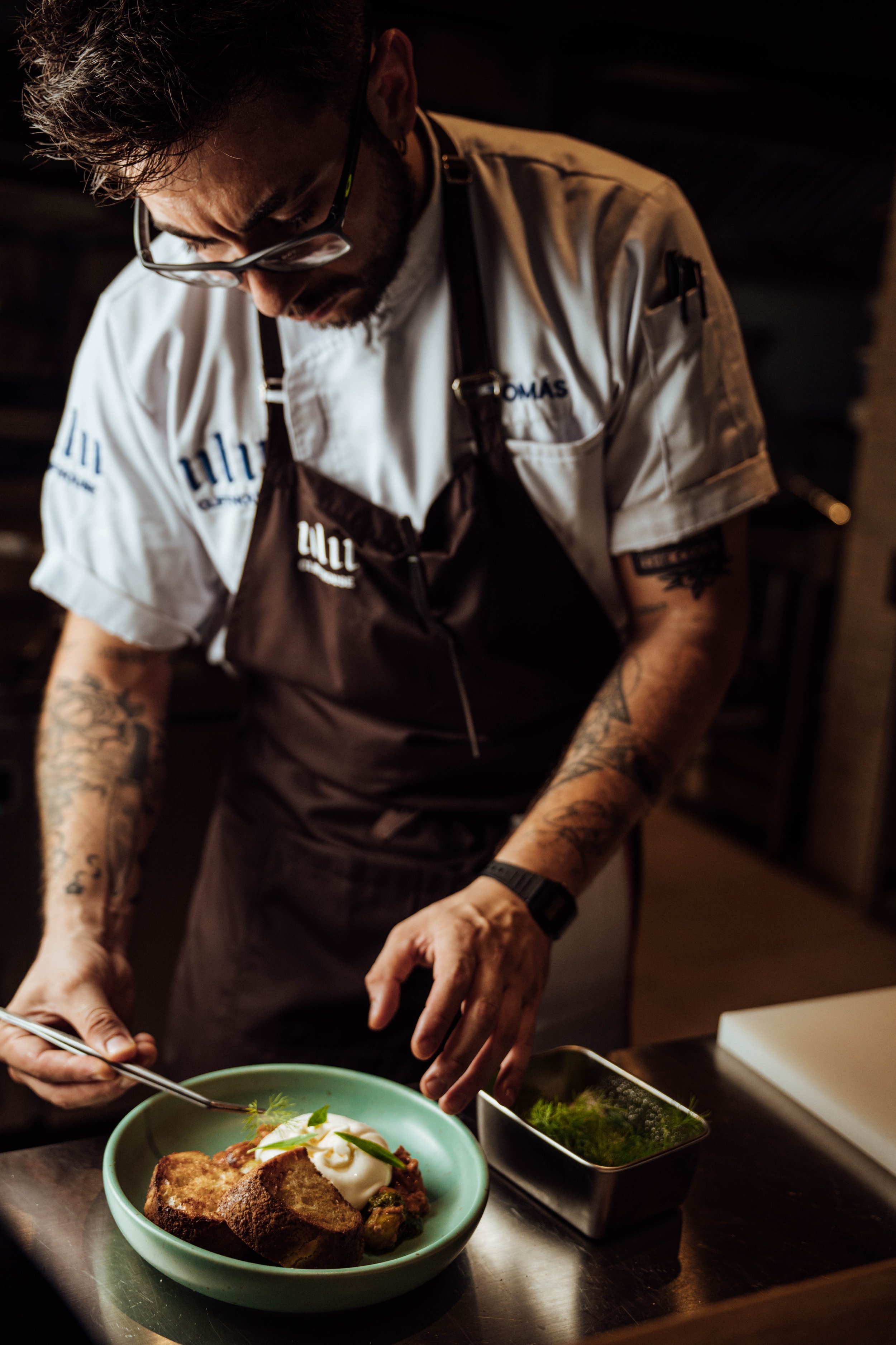 A chef wearing glasses and a white shirt with various logos, including 'TOMAS', is preparing a dish in a kitchen. The dish contains pieces of bread, what looks like poached eggs, and some greens in a bowl. There are additional ingredients in small containers nearby.