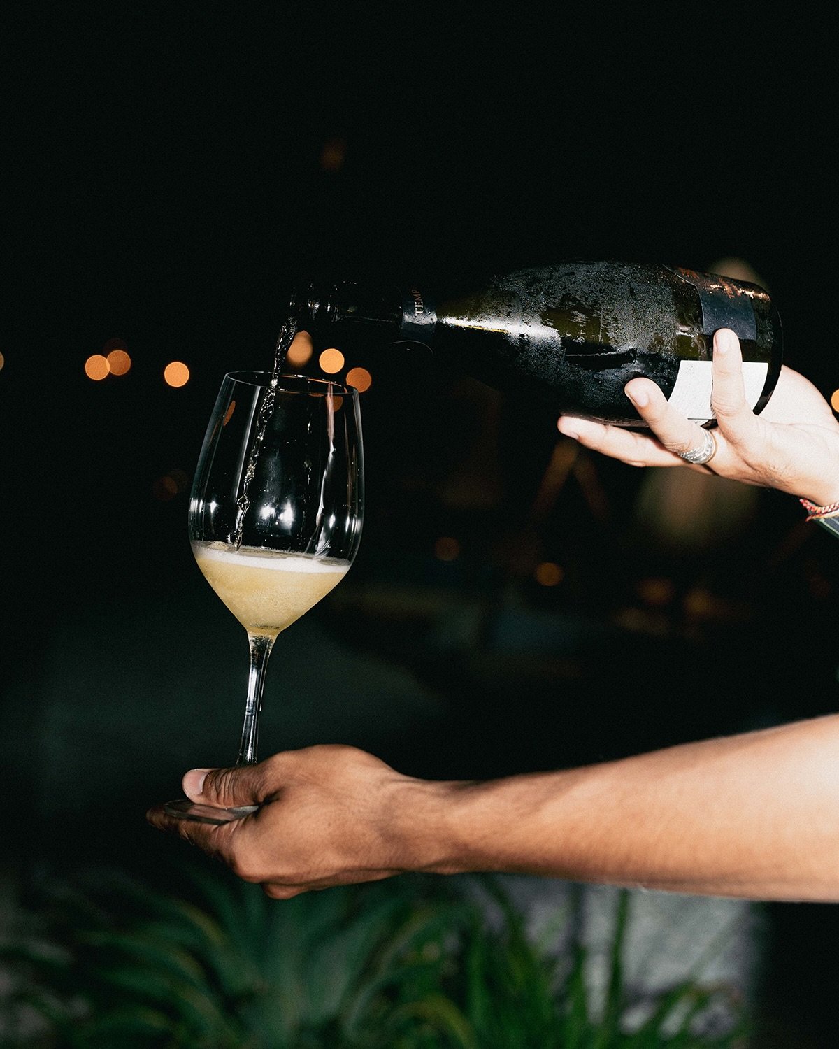 Person pouring champagne into a wine glass at night with blurred lights in the background.