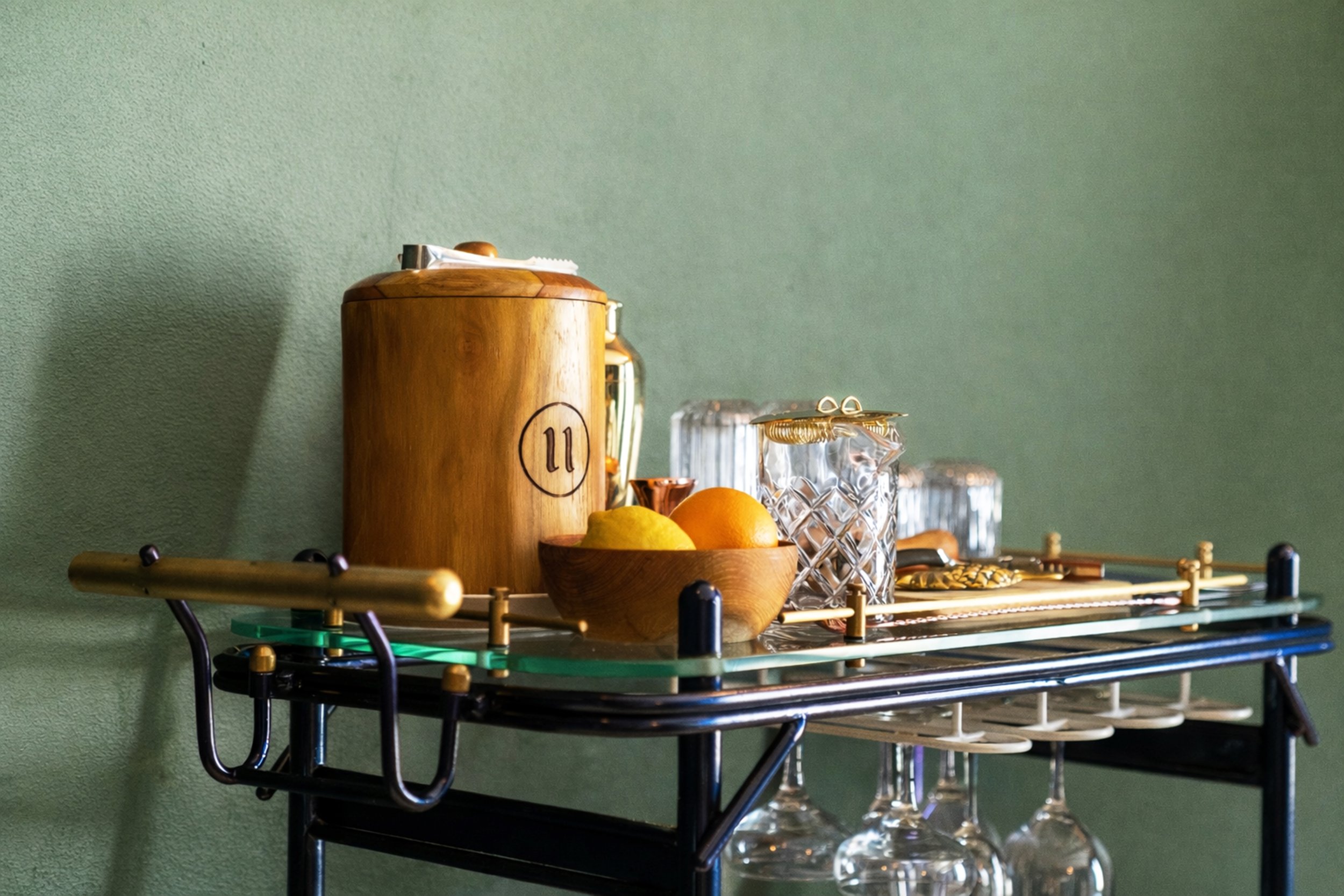 A mid-century modern wooden container with a lid, placed on a table decorated with various glassware, fruits, and a miniature golf set against a green textured wall.