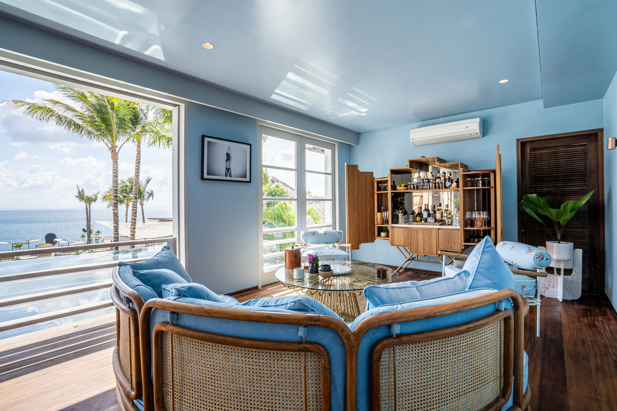 Cozy living room with large windows overlooking a beach scene with palm trees, blue sky, and ocean, featuring blue cushioned rattan furniture, a wooden bar, and beach-themed decor.