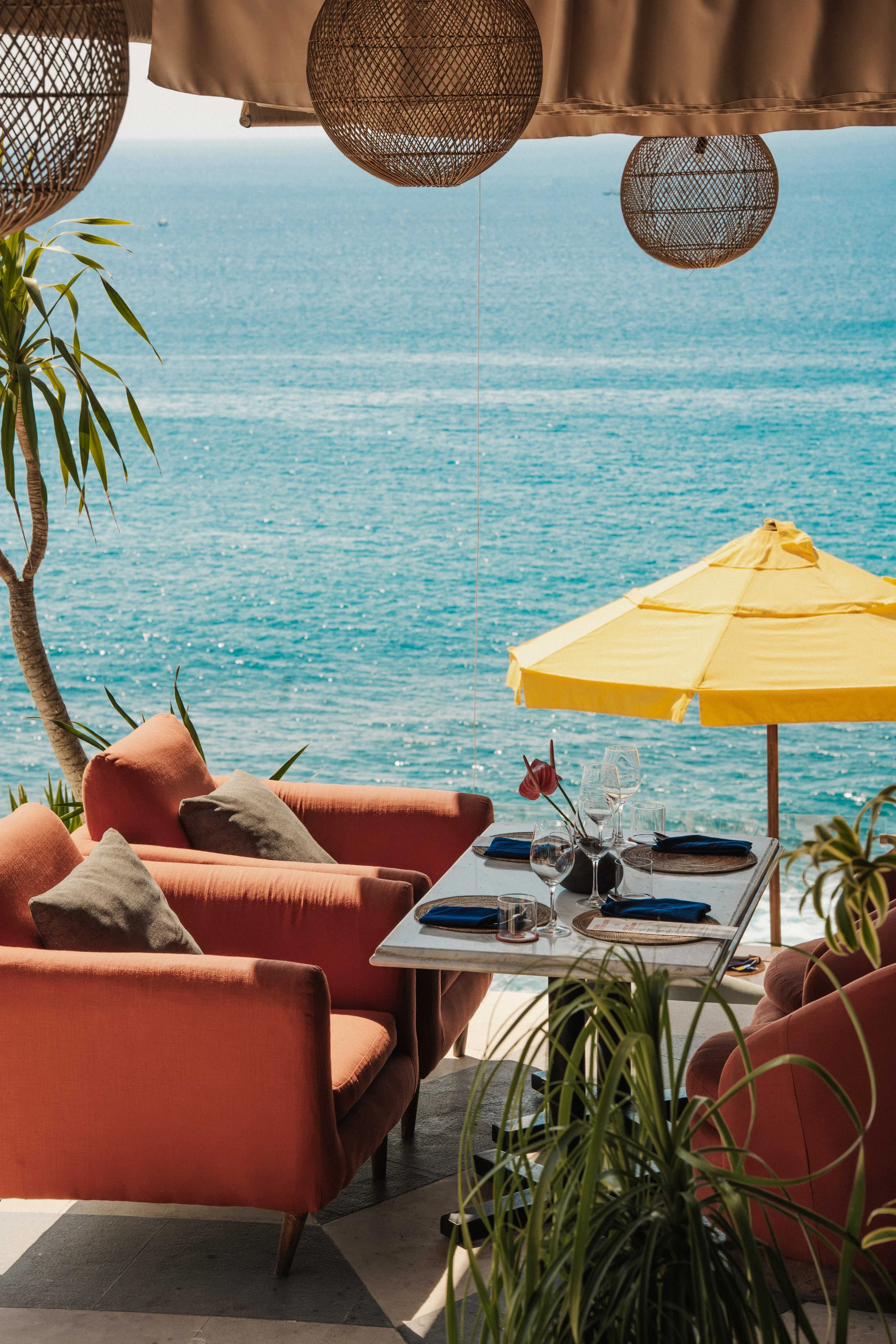 A seaside outdoor dining area with pink sofas, a table set with glassware and napkins, a yellow umbrella, and decorative hanging lamps under a canopy, overlooking the ocean.