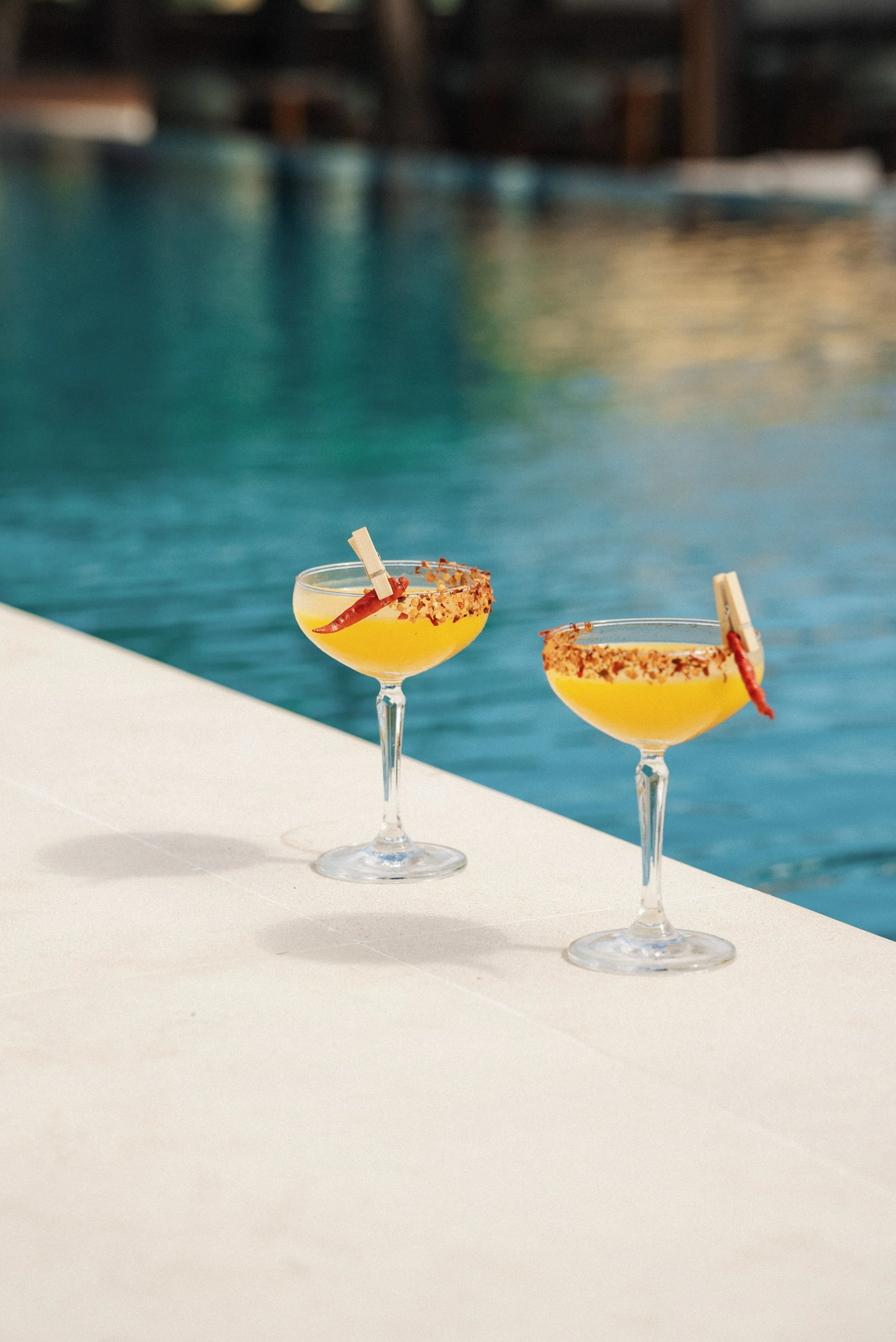 Two cocktails with yellow liquid, garnished with chili peppers and salt, on a white surface near a swimming pool.