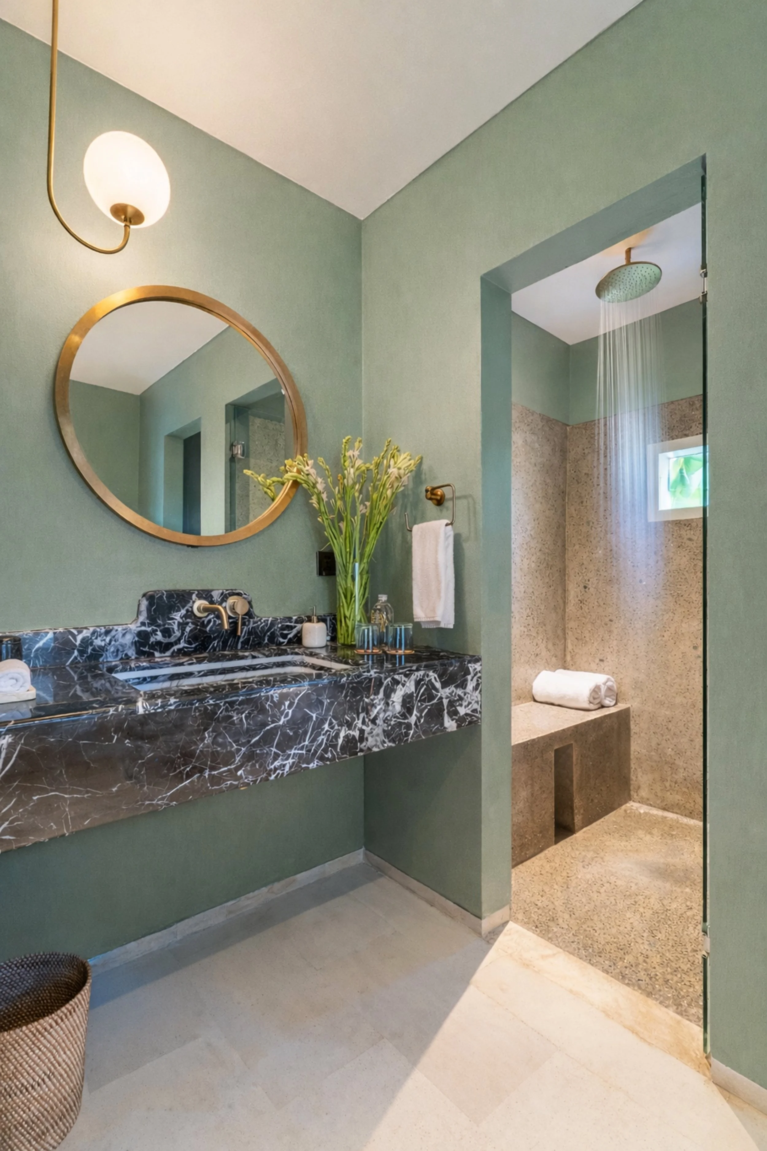 A modern bathroom with a green wall, a black marble sink with gold fixtures, a round mirror, a vase with flowers, bathroom essentials, and a separate shower area with a rainfall showerhead and beige tiling.