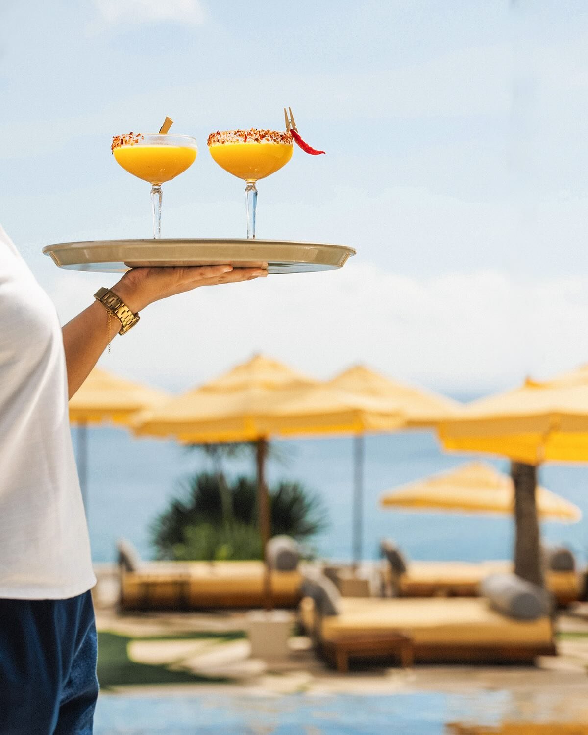 A person holding a tray with two yellow drinks in coupe glasses with garnishes, against a background of beach umbrellas and chairs at the beach.