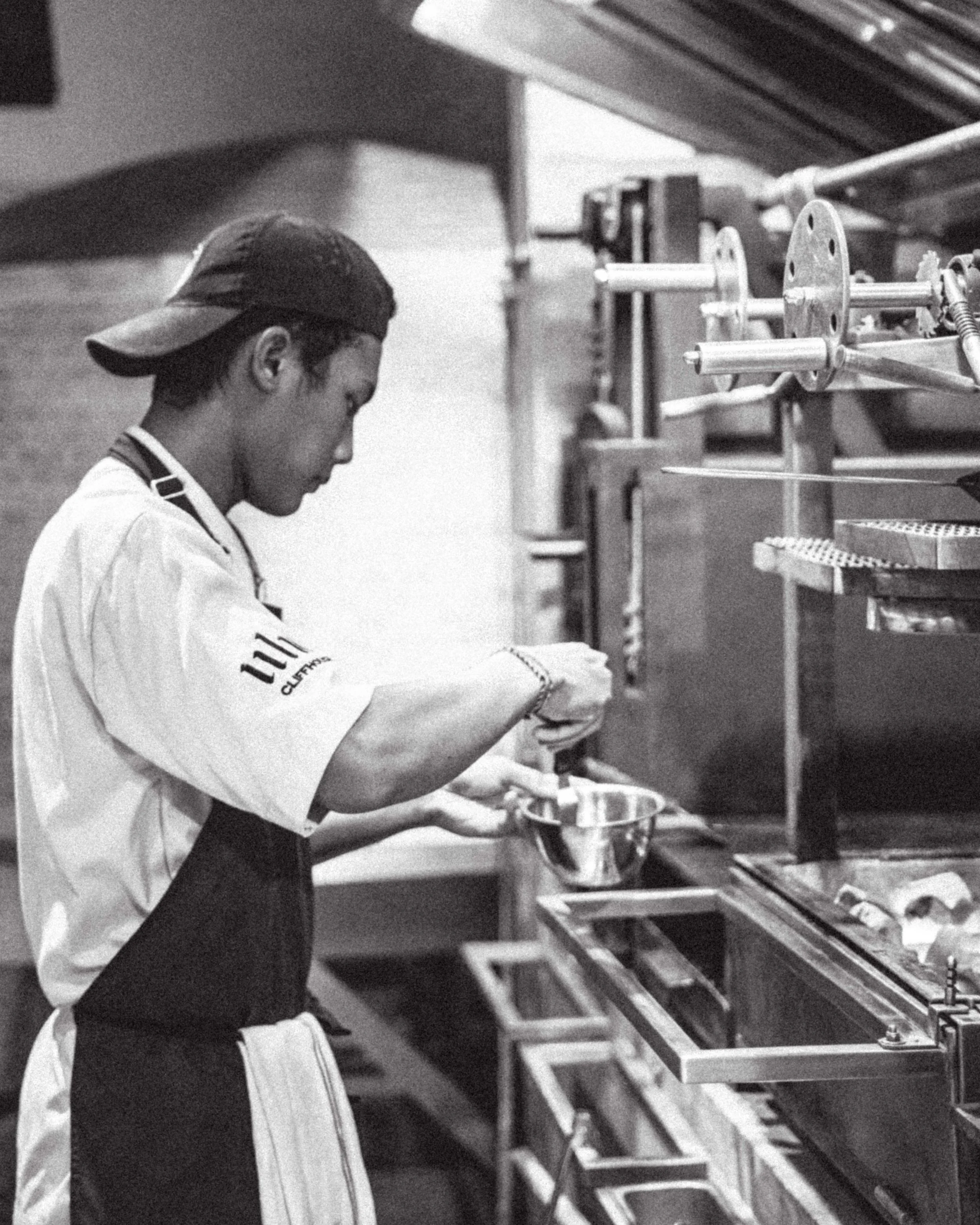 Black and white photo of a person in a kitchen or bakery, wearing a cap and apron, working at a countertop with industrial baking or cooking equipment.