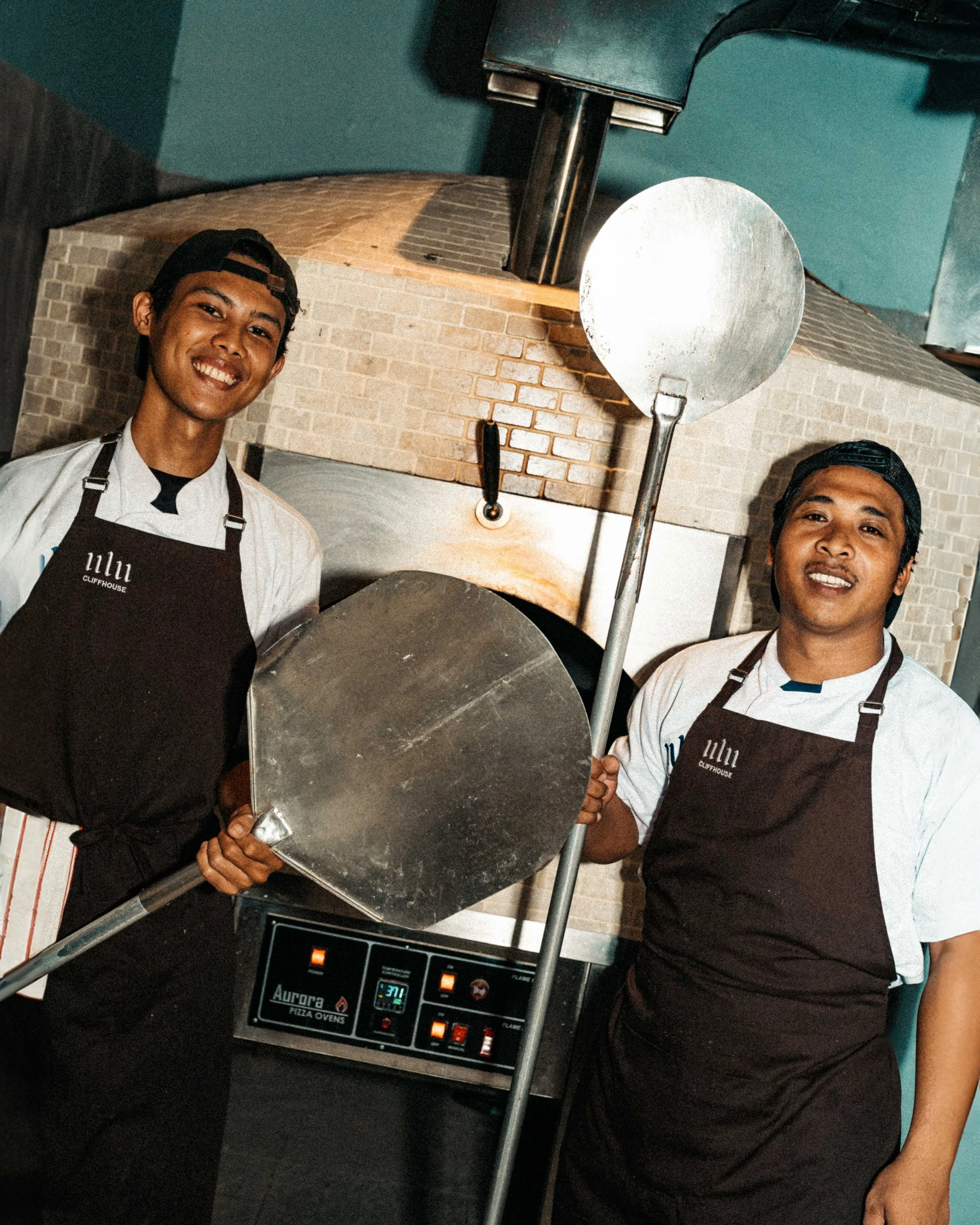 Two chefs in white uniforms and black aprons, with one wearing a black cap, smiling in a kitchen with a brick wall background, with a pizza oven and a digital control panel in the foreground.