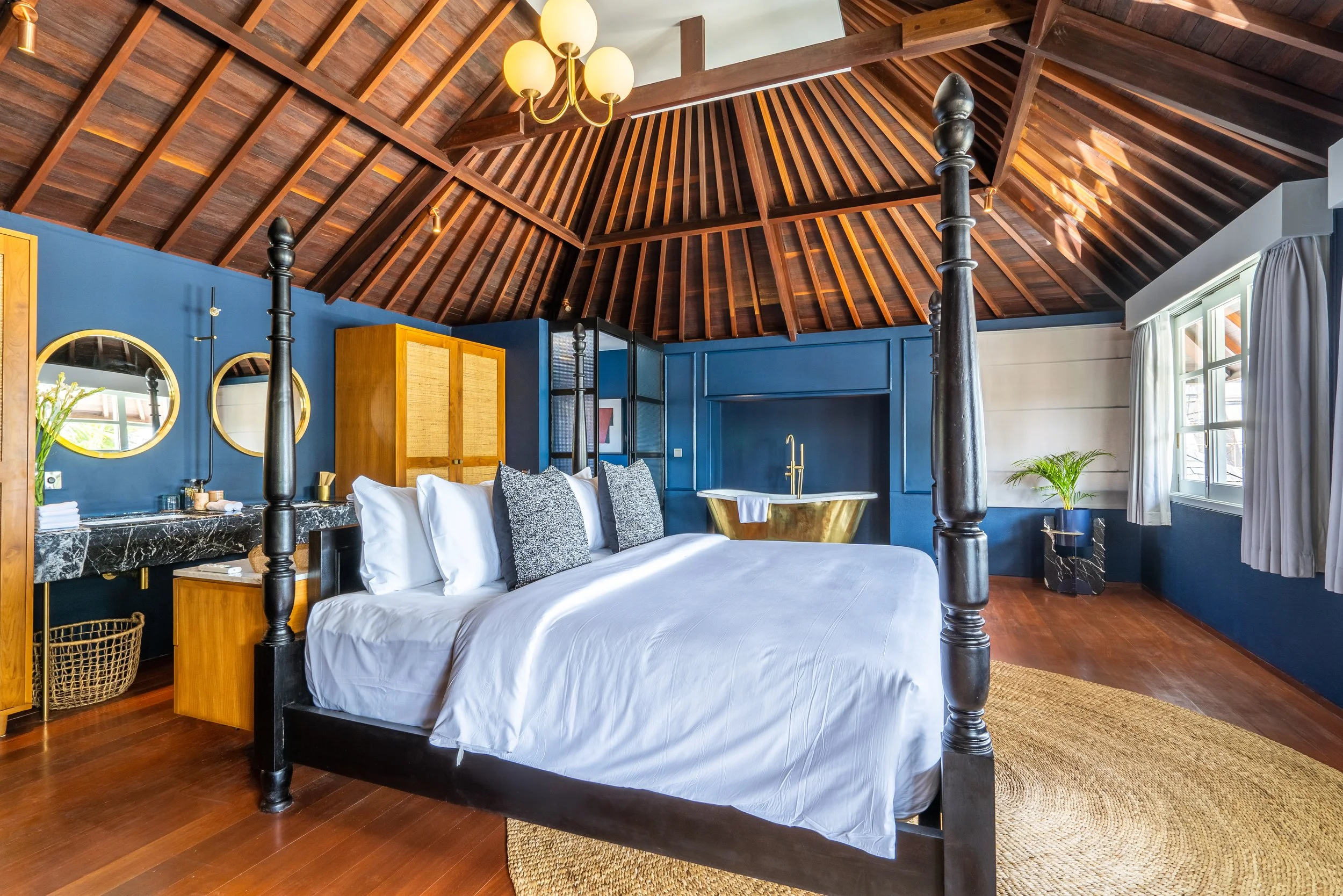 A bedroom with a dark wooden four-poster bed, white bedding, and gray pillows. The room features a vaulted wooden ceiling with a chandelier, blue accent walls, two round mirrors, a black marble vanity with gold accents, a brass freestanding bathtub, 