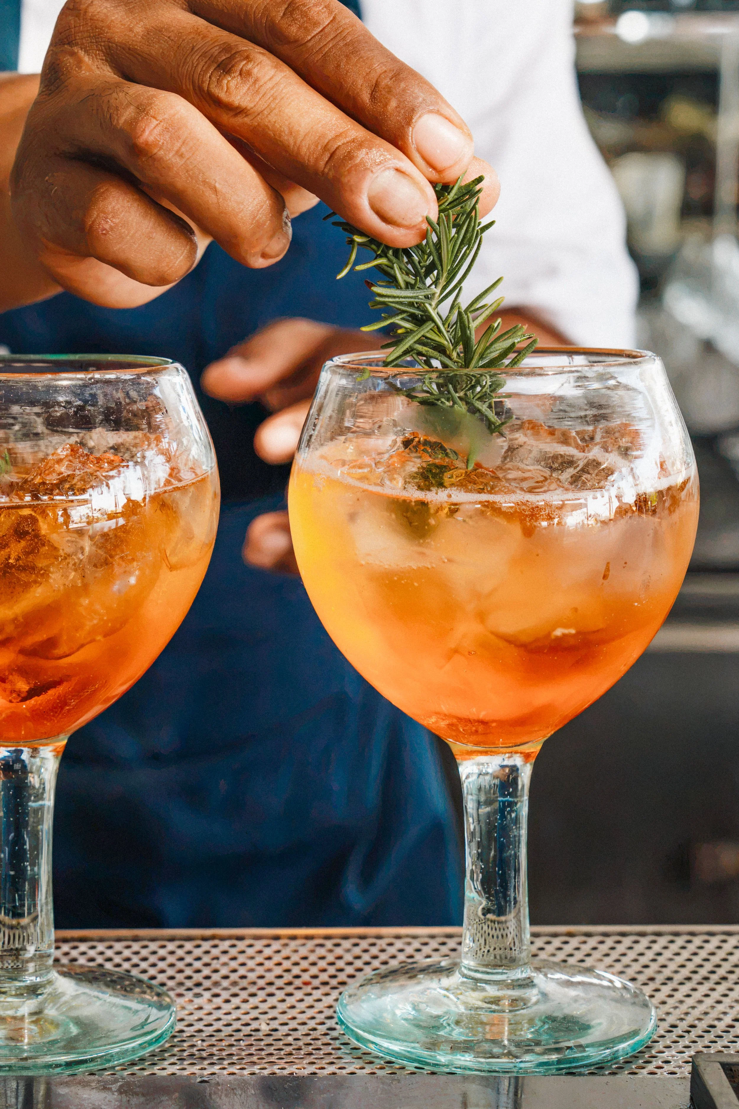 A person garnishing two glasses of orange-colored cocktail with a sprig of rosemary.
