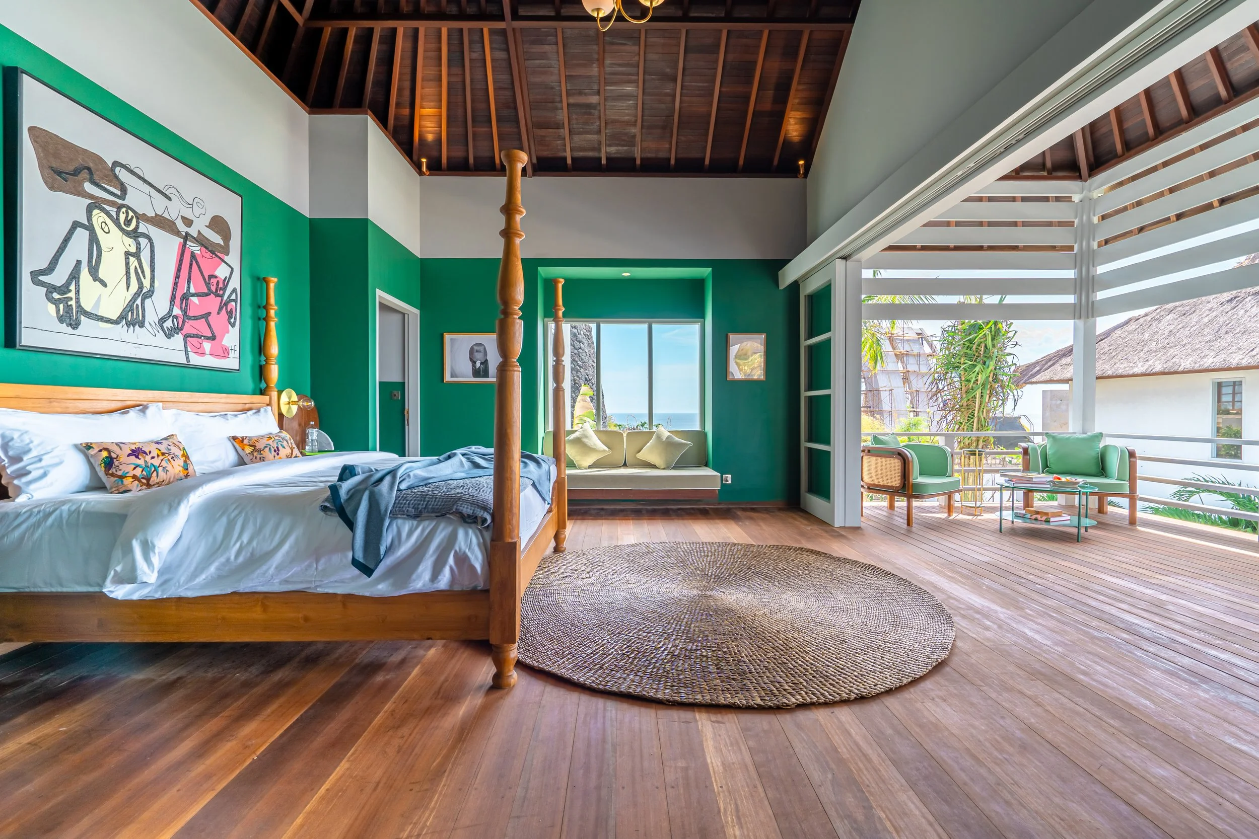 A spacious tropical hotel room with a wooden four-poster bed, colorful floral pillows, and artwork on the walls. Large glass doors lead to a balcony with seating and views of palm trees and thatched roofs. The room features a high, wooden vaulted cei